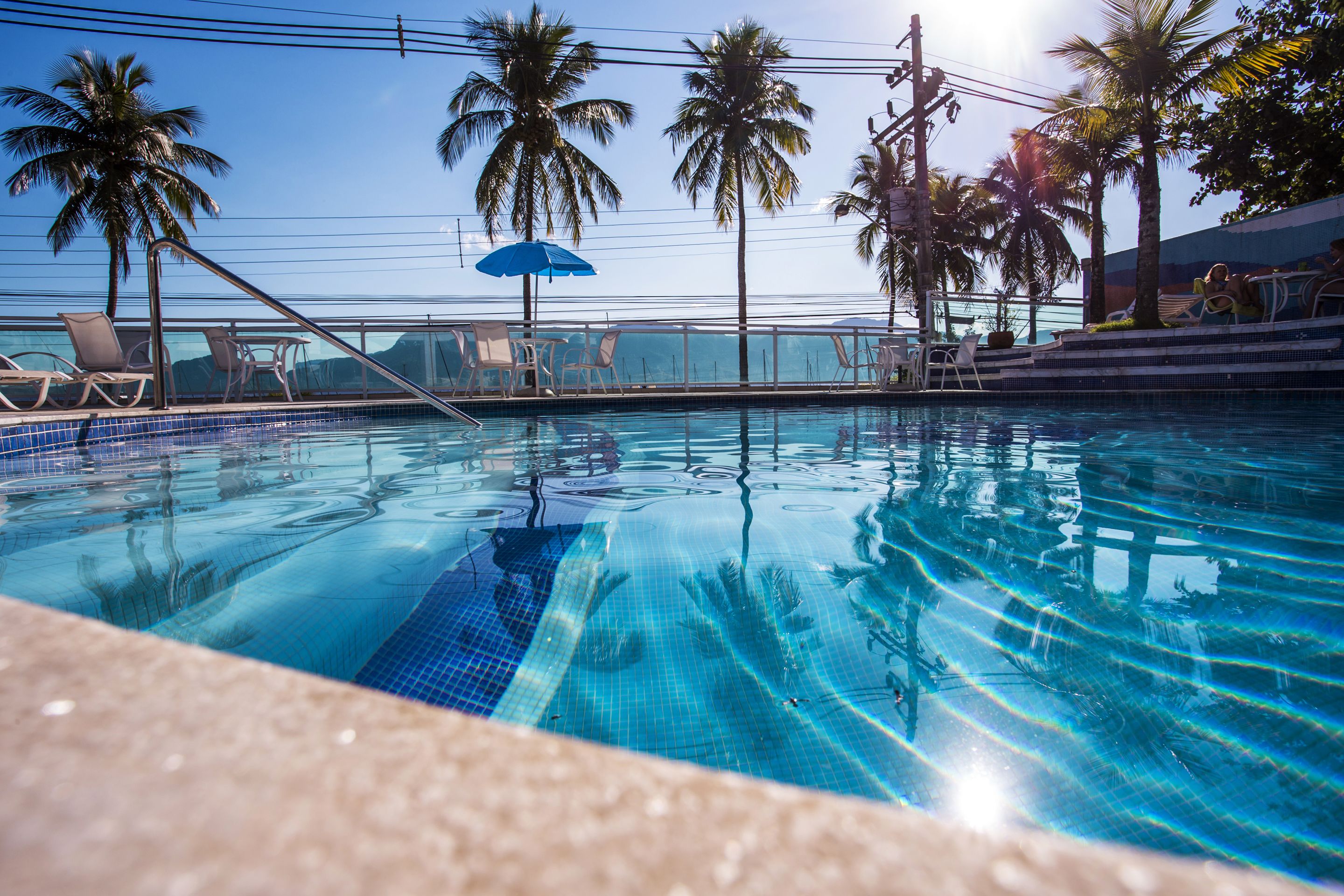 Una piscina azul cristalina con sillas de descanso y sombrillas, rodeada de palmeras.
