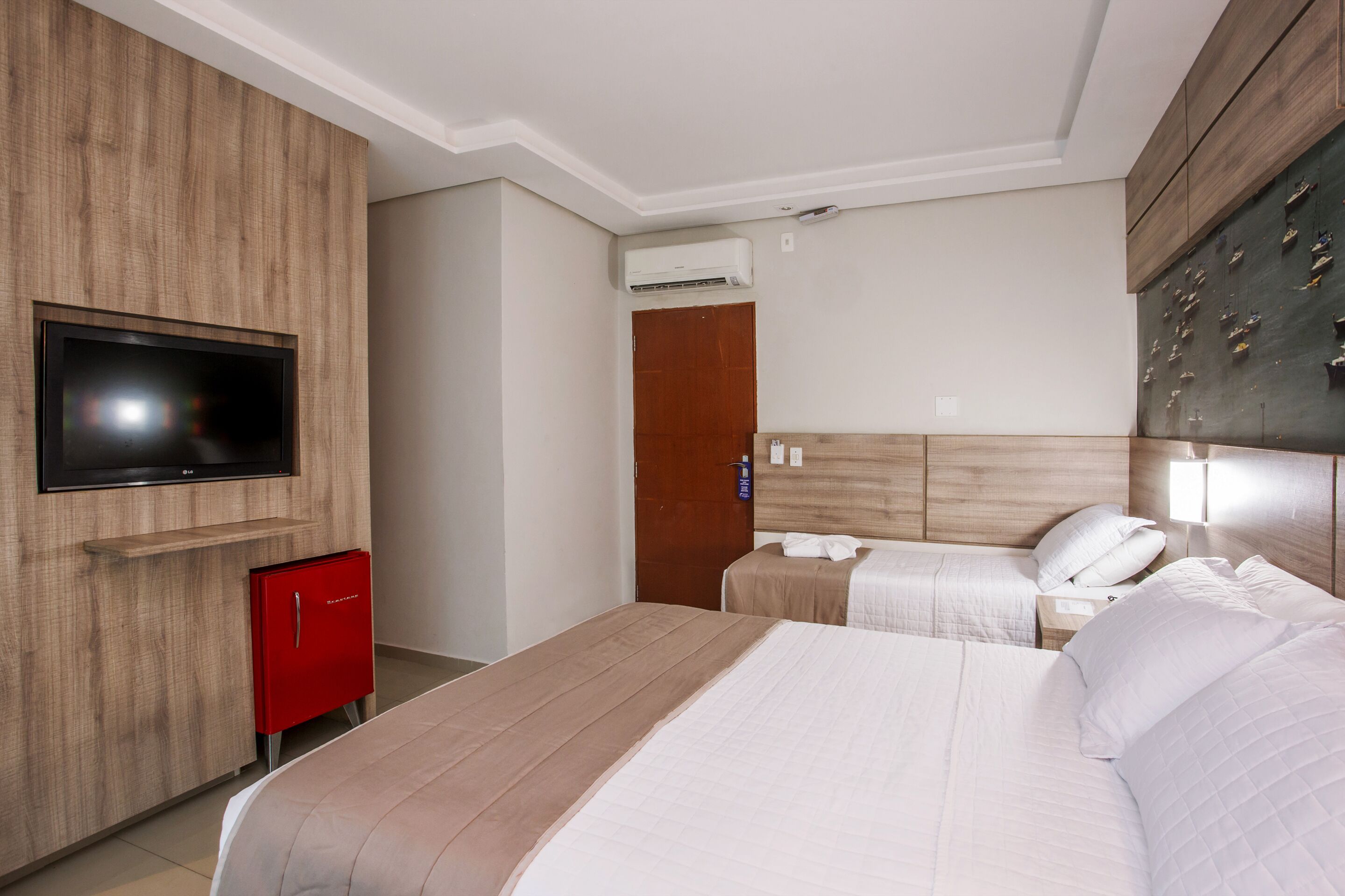 Una habitación de hotel con una cama grande, un televisor montado en la pared, un minibar rojo y un cabecero de madera.