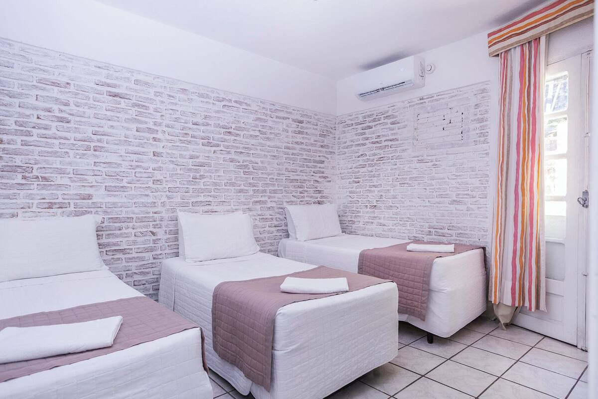 Una habitación con tres camas, ropa de cama blanca, una pared con diseño de ladrillo y una cortina a rayas.