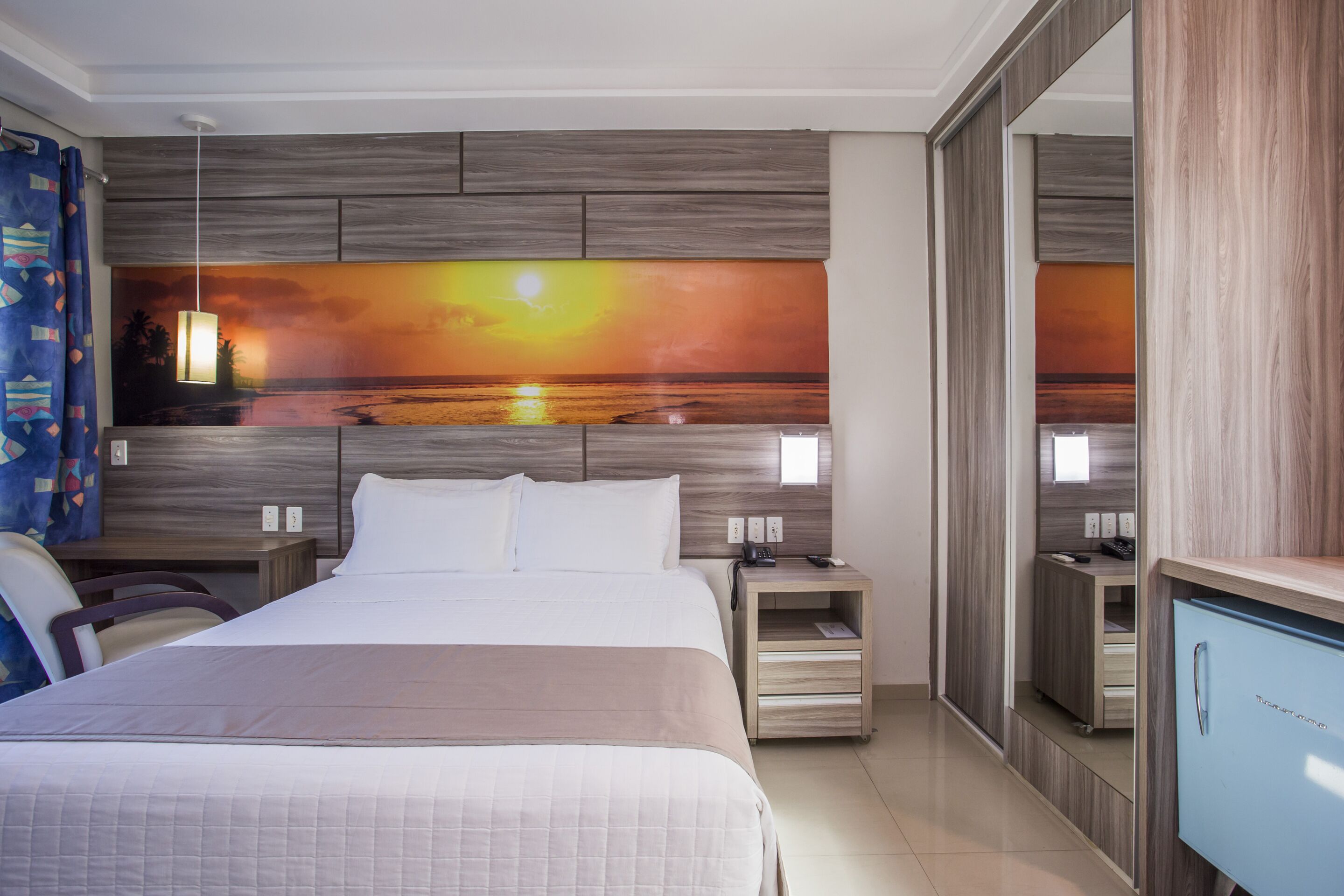 Quarto de hotel com uma cama grande, mesa de cabeceira, escrivaninha, espelho e guarda-roupa.
