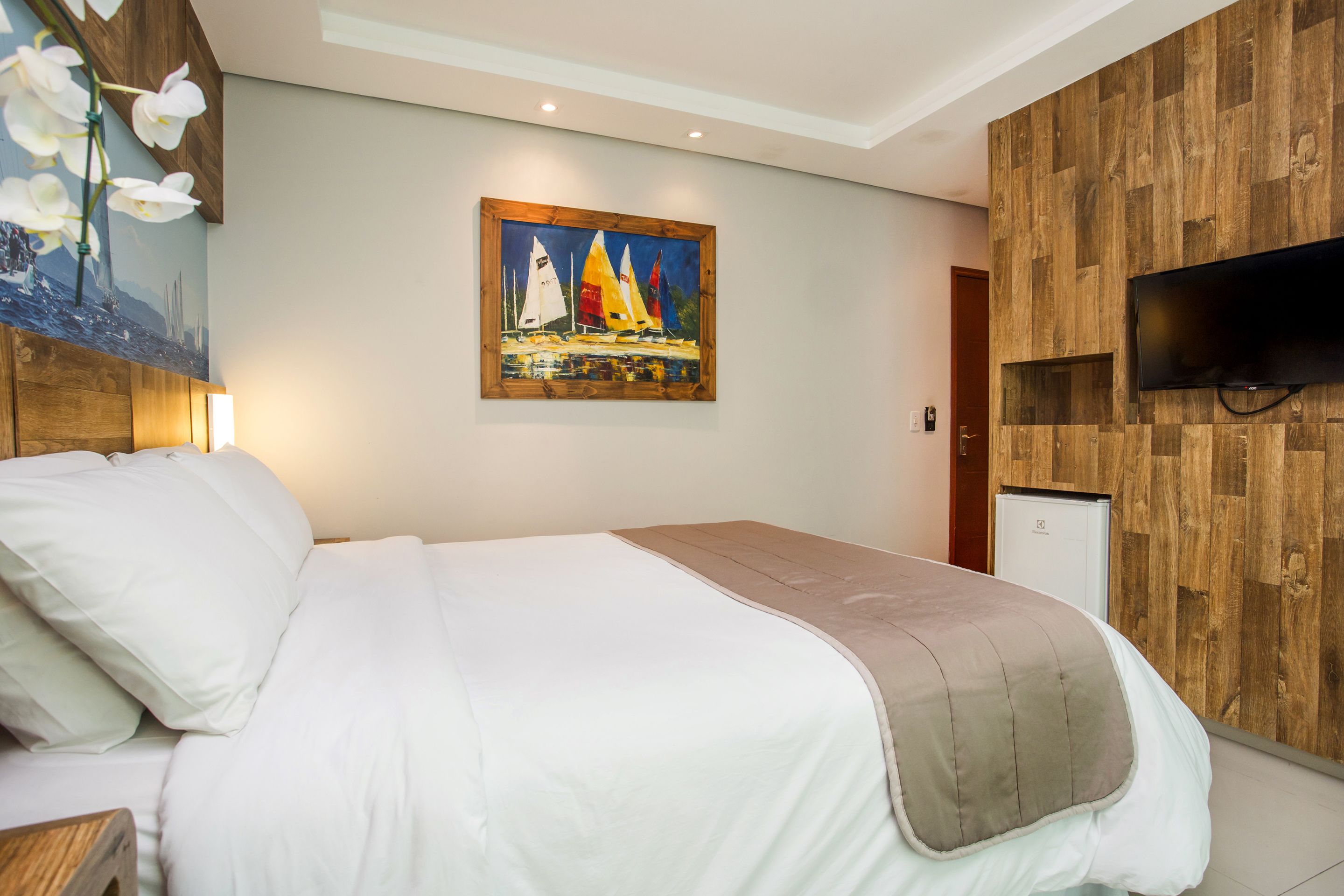 A hotel room with a bed, a painting of sailboats, a television, and a wooden wall panel.