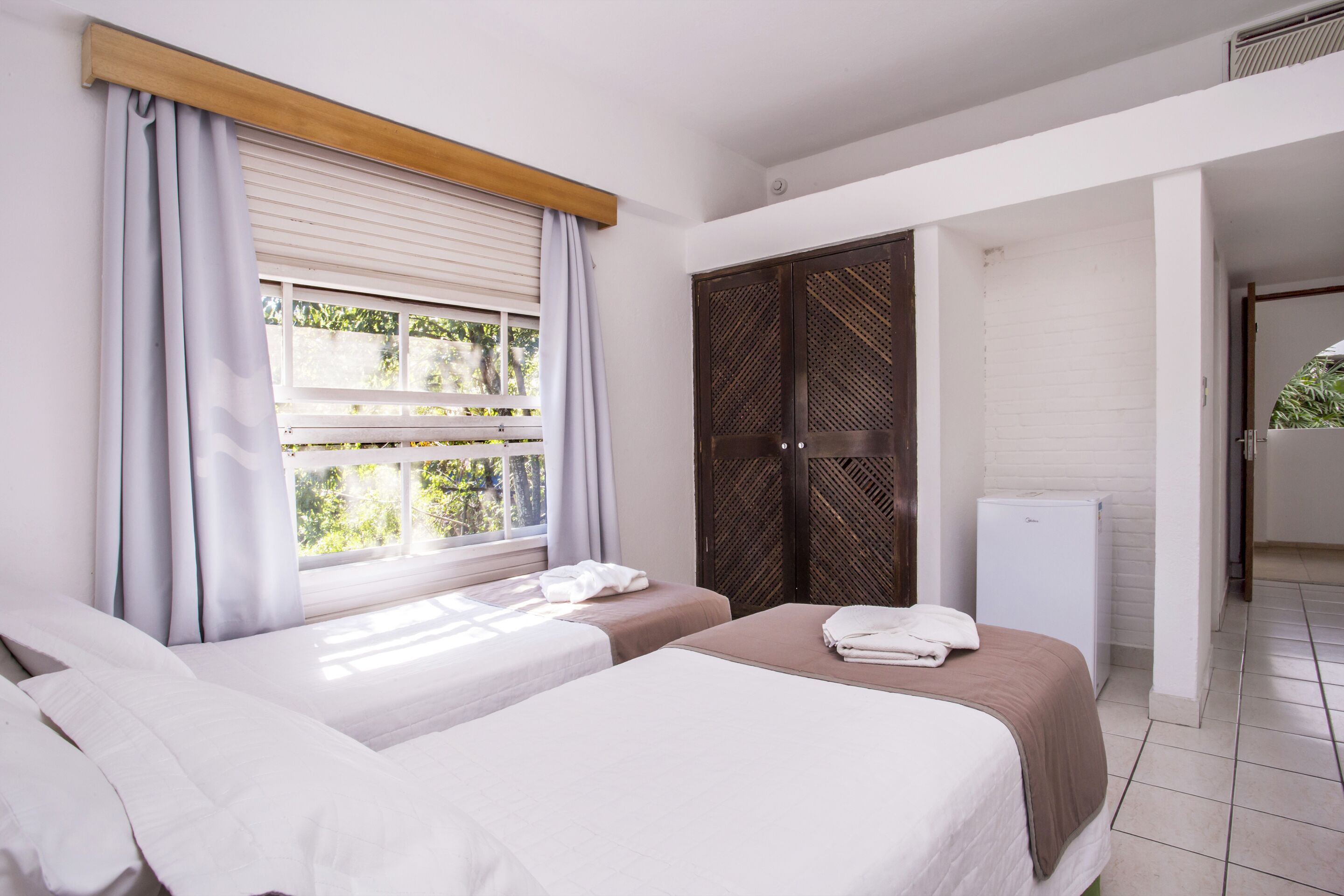 Een hotelkamer met twee bedden, een kledingkast, een kleine koelkast en een raam met gordijnen.