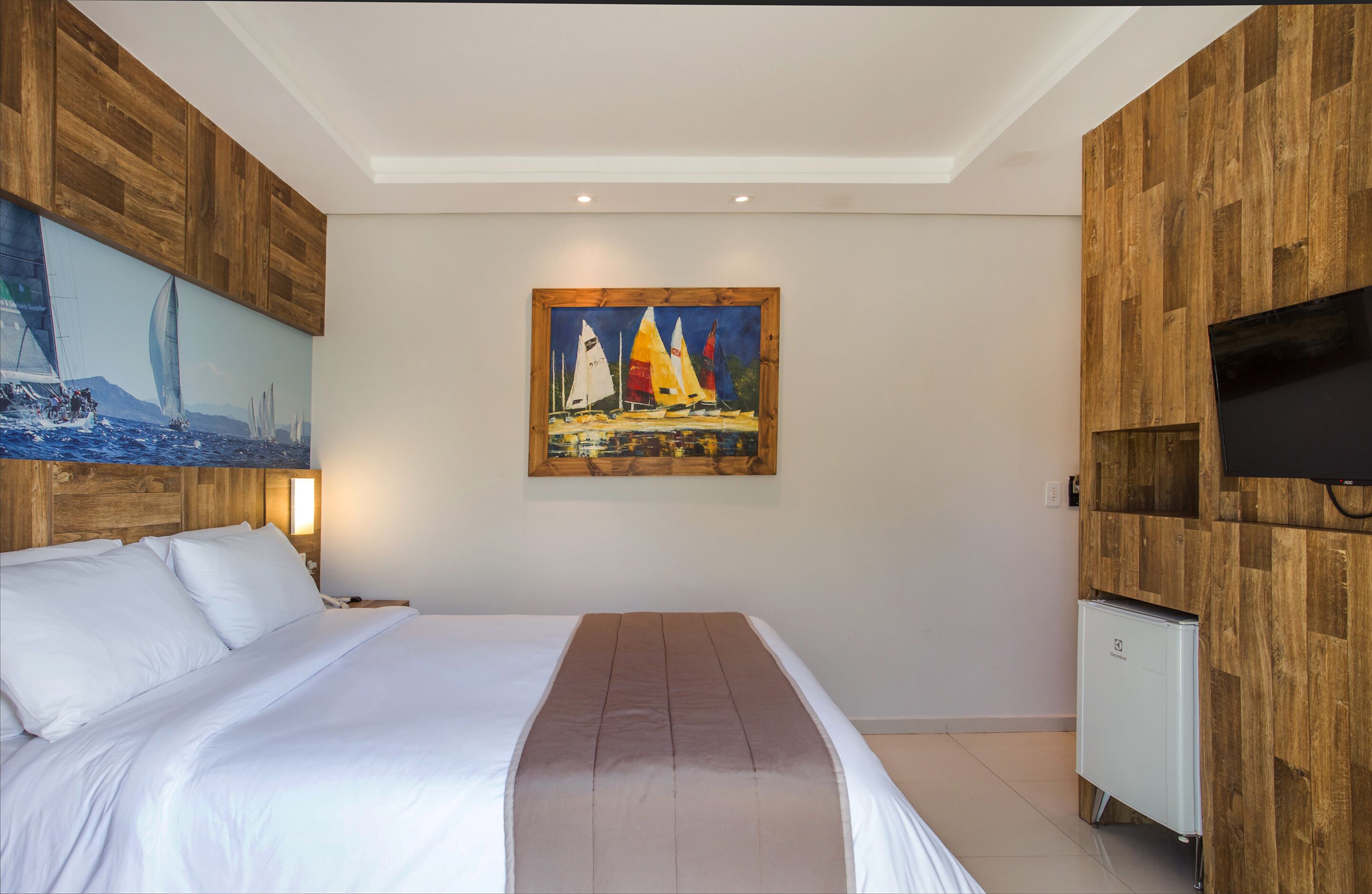 Un dormitorio con una cama grande, un cuadro de veleros, un televisor y un minibár.