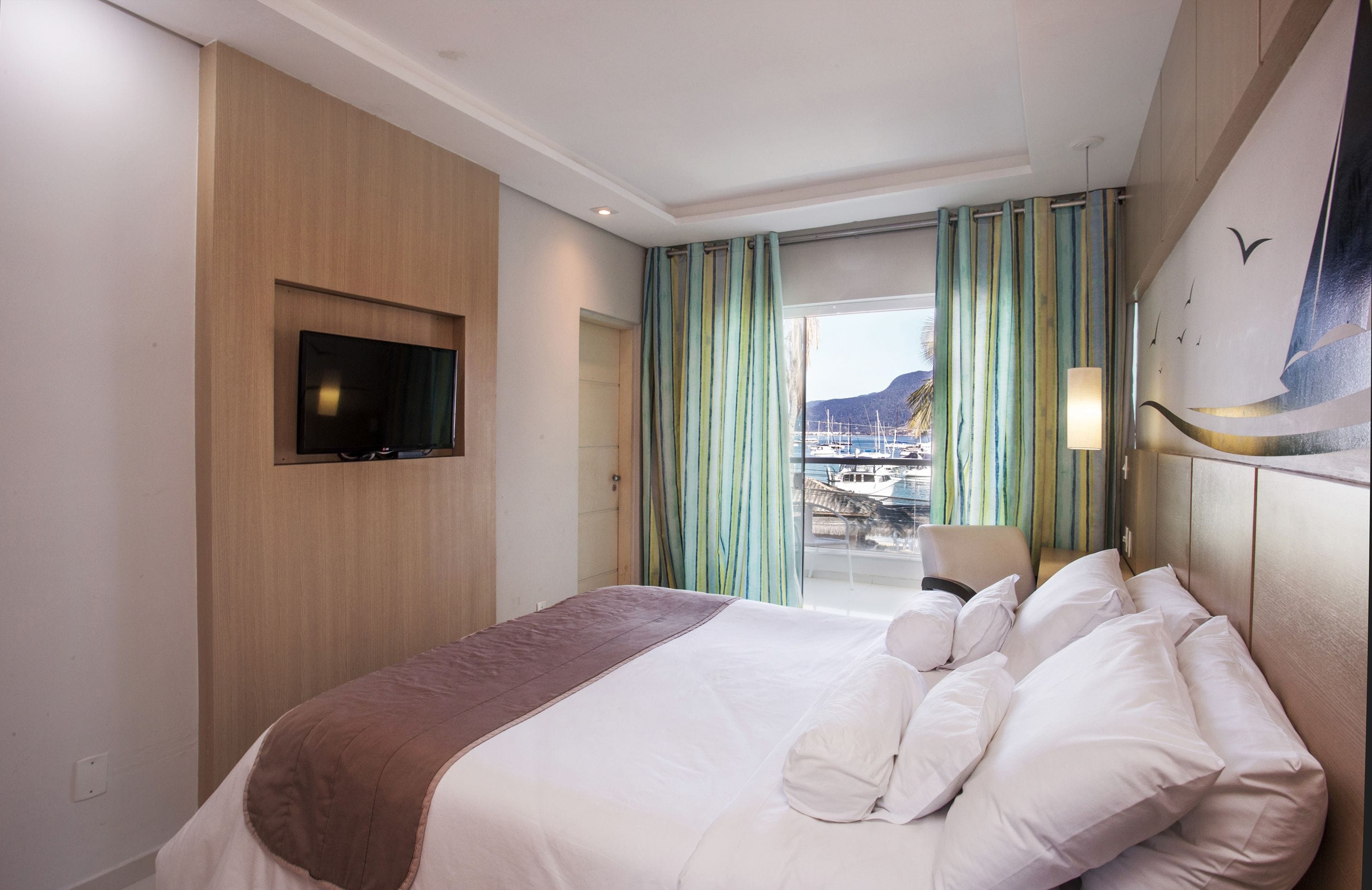 Ein Hotelzimmer mit Bett, Wandfernseher, Blick auf einen Yachthafen mit Booten und einem Balkon mit grünen Vorhängen.