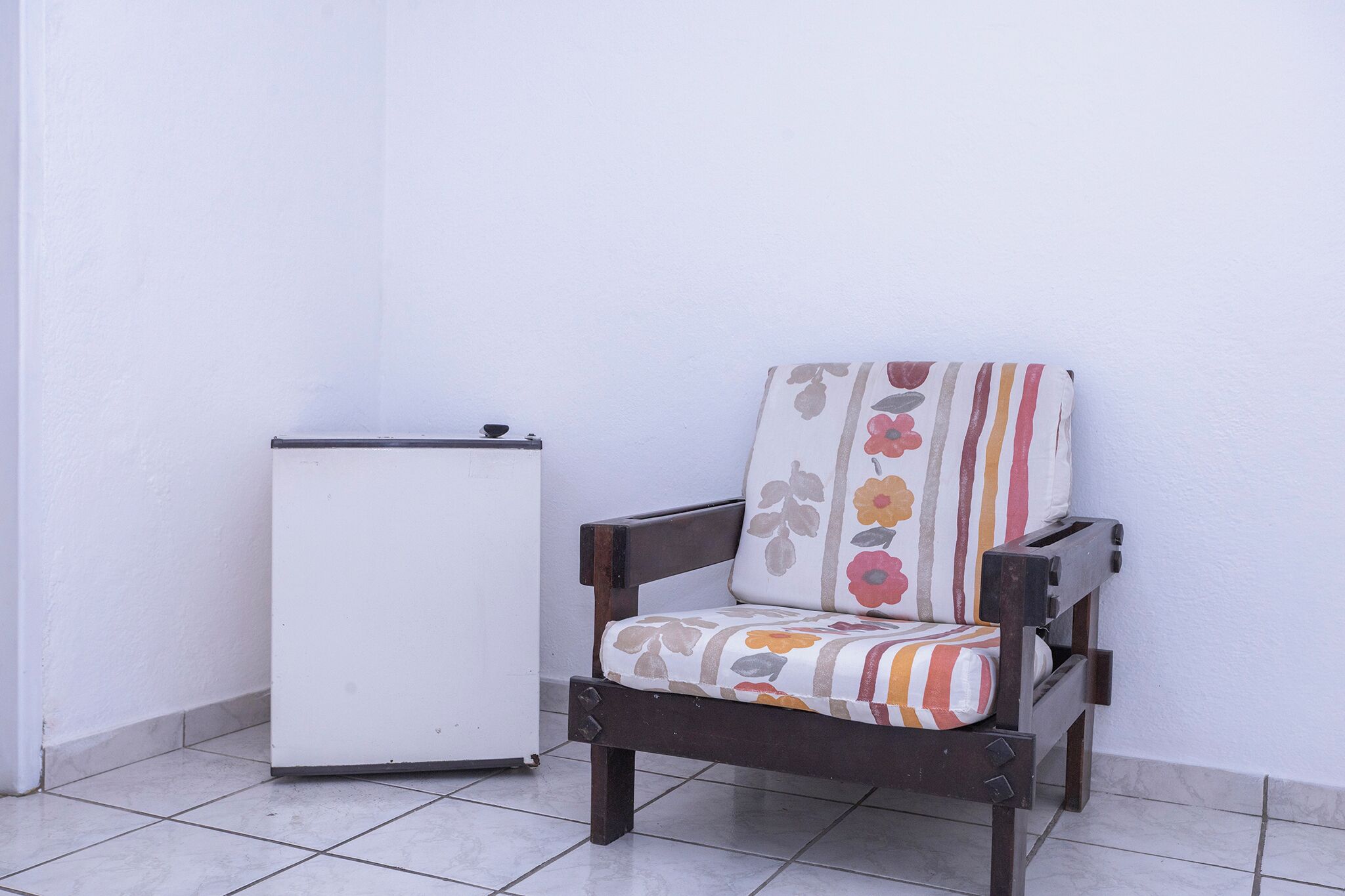 A corner of a room with a white wall and a tiled floor. There is a small white refrigerator and a wooden chair with a floral cushion.