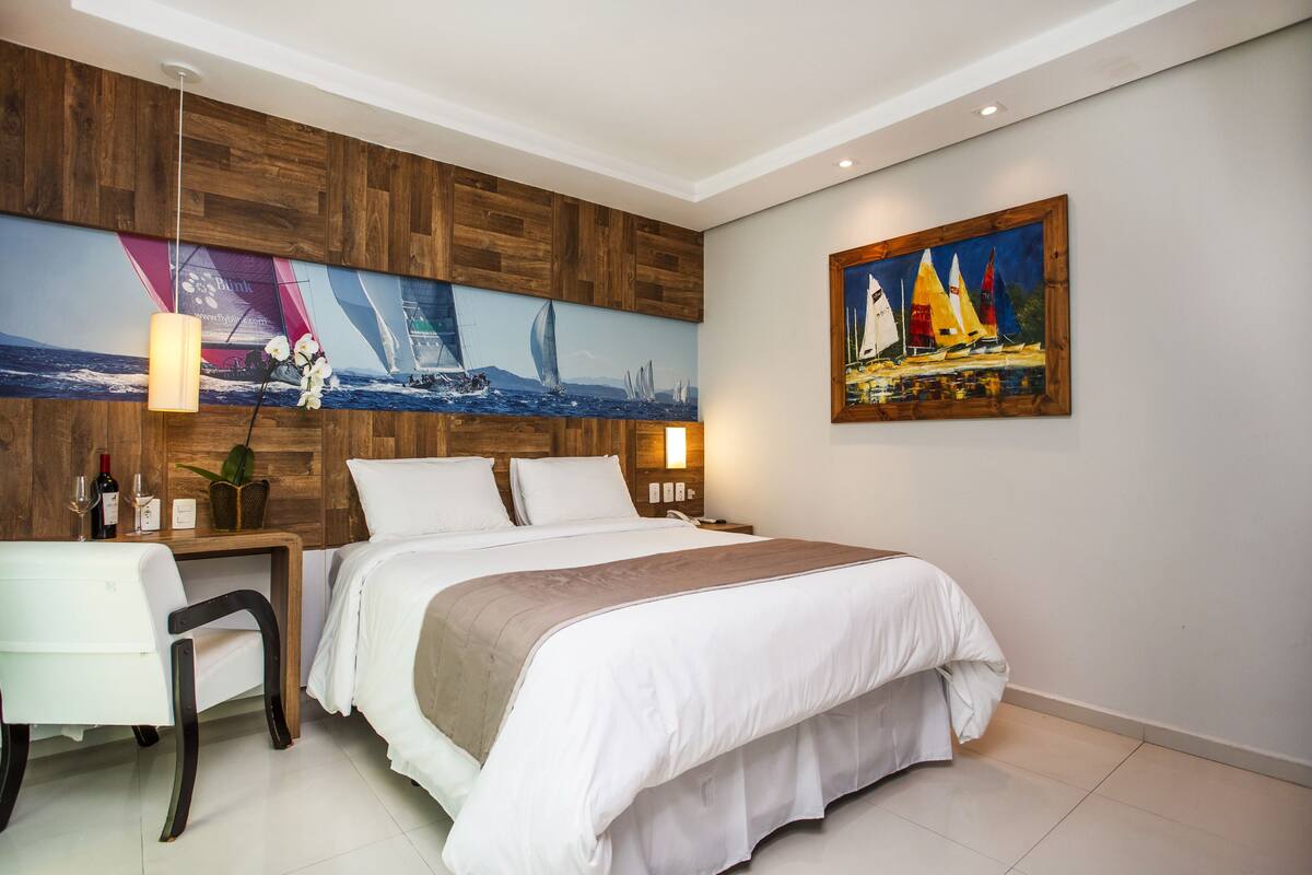 Una habitación de hotel moderna con una cama grande, un escritorio, una silla, un panel de madera decorativo y una pintura de veleros.