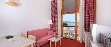 A compact room with a sofa, a small table with a fruit bowl, a TV, and a balcony with a view.