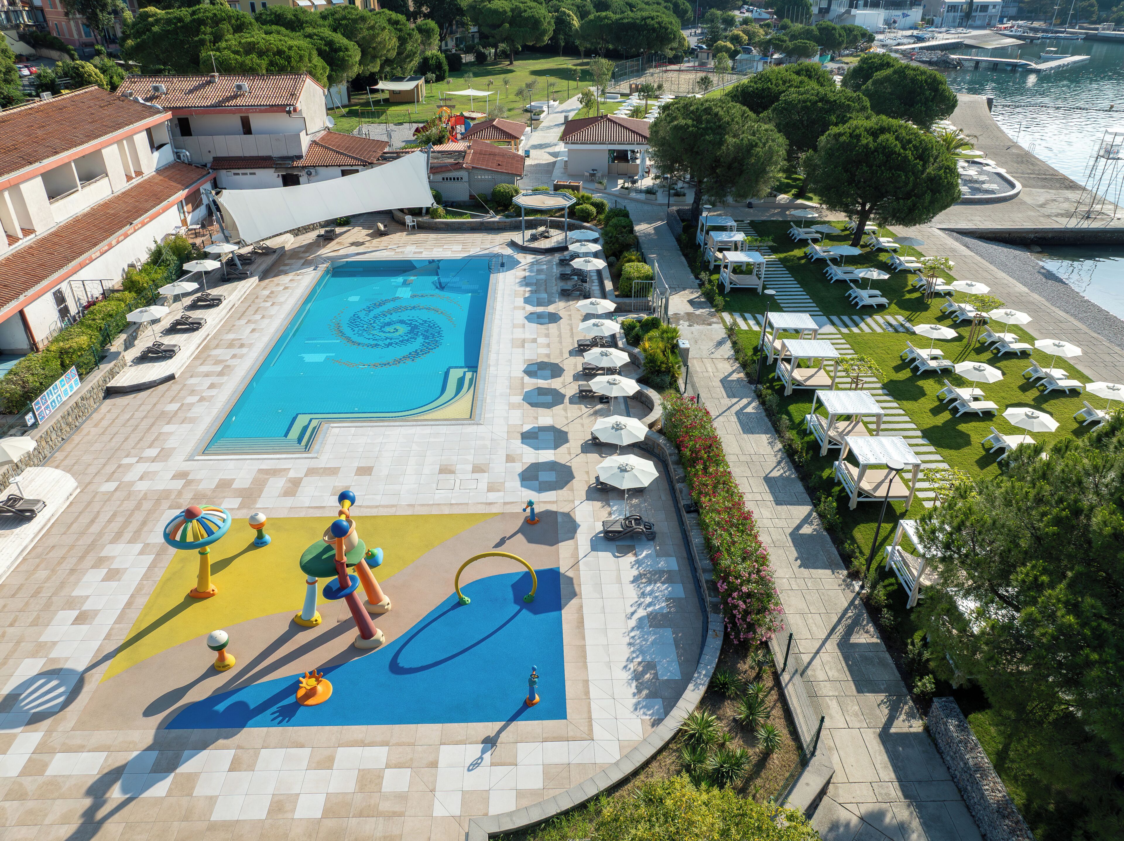 A swimming pool with a spiral design, surrounded by lounge chairs and umbrellas, with a view of the sea and buildings in the background.
