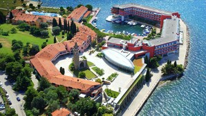 Vista aérea de un hotel frente al mar con piscina, un edificio grande de fachada roja y un puerto deportivo con barcos.