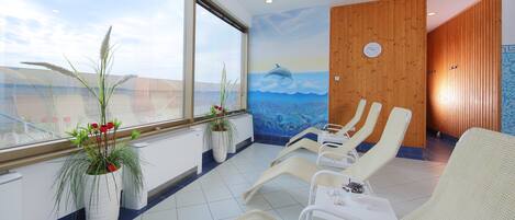 A sunlit room with a large window, a wooden wall with a clock, and several lounge chairs.