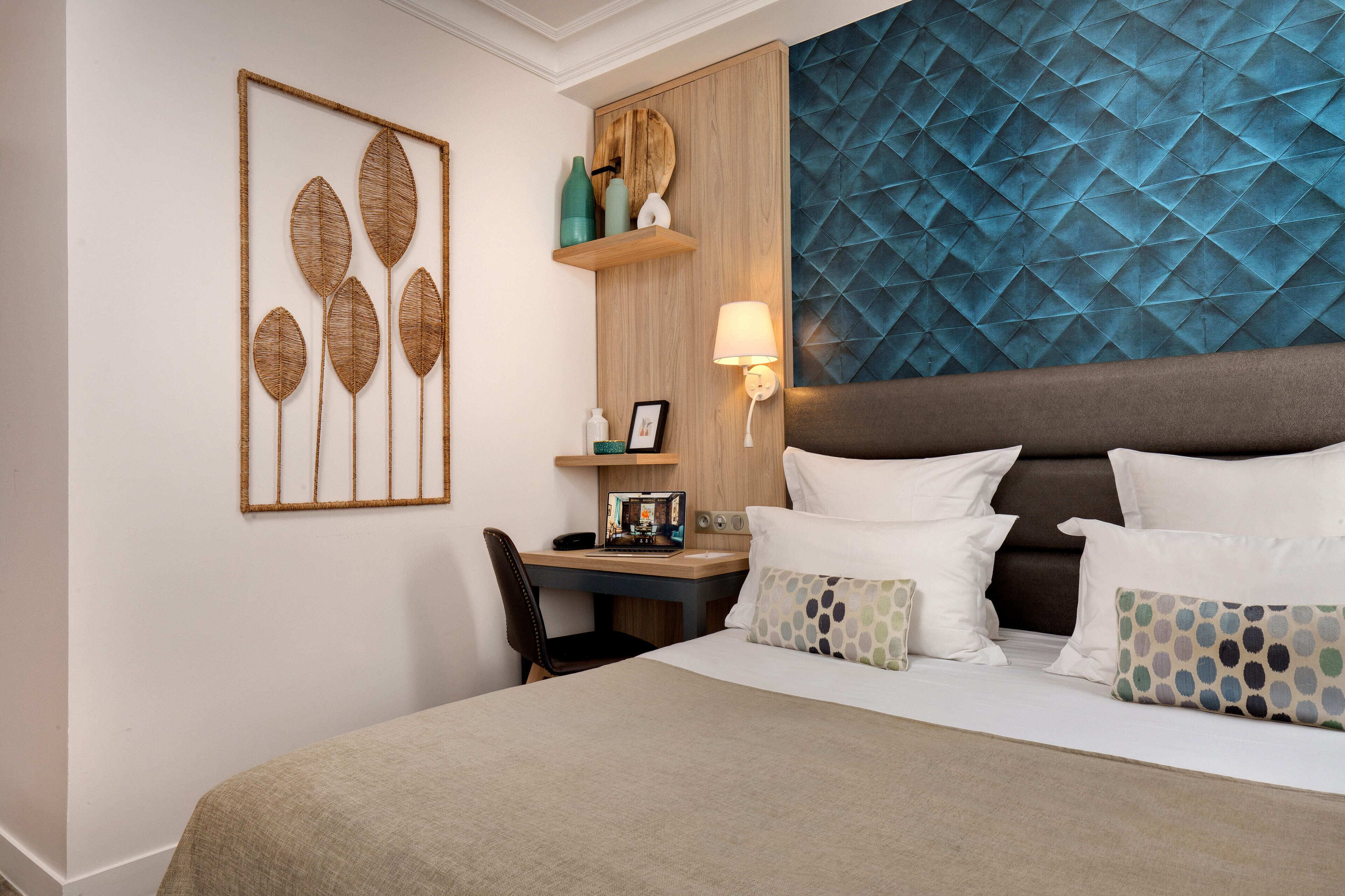 A hotel room with a bed, a desk, a chair, a wall-mounted shelf with decorative items, and a wall with a geometric pattern.