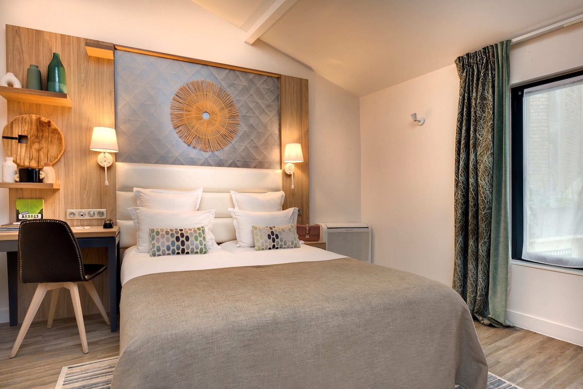 Une chambre d’hôtel avec un grand lit, un bureau et une chaise. Un étagère fixée au mur est décorée de divers objets, et une grande œuvre d’art circulaire est accrochée au-dessus du lit.