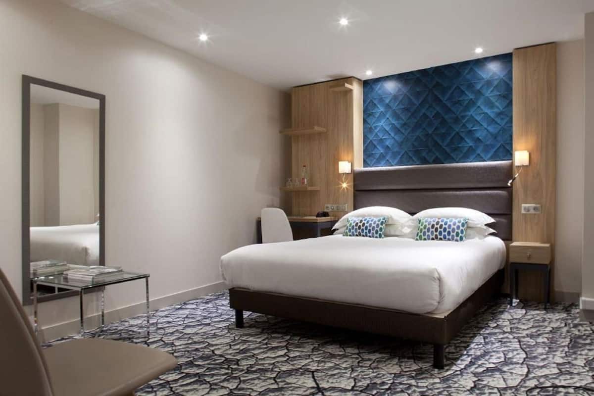 Une chambre d’hôtel avec un lit, un bureau, une chaise, un miroir et un sol à motifs.