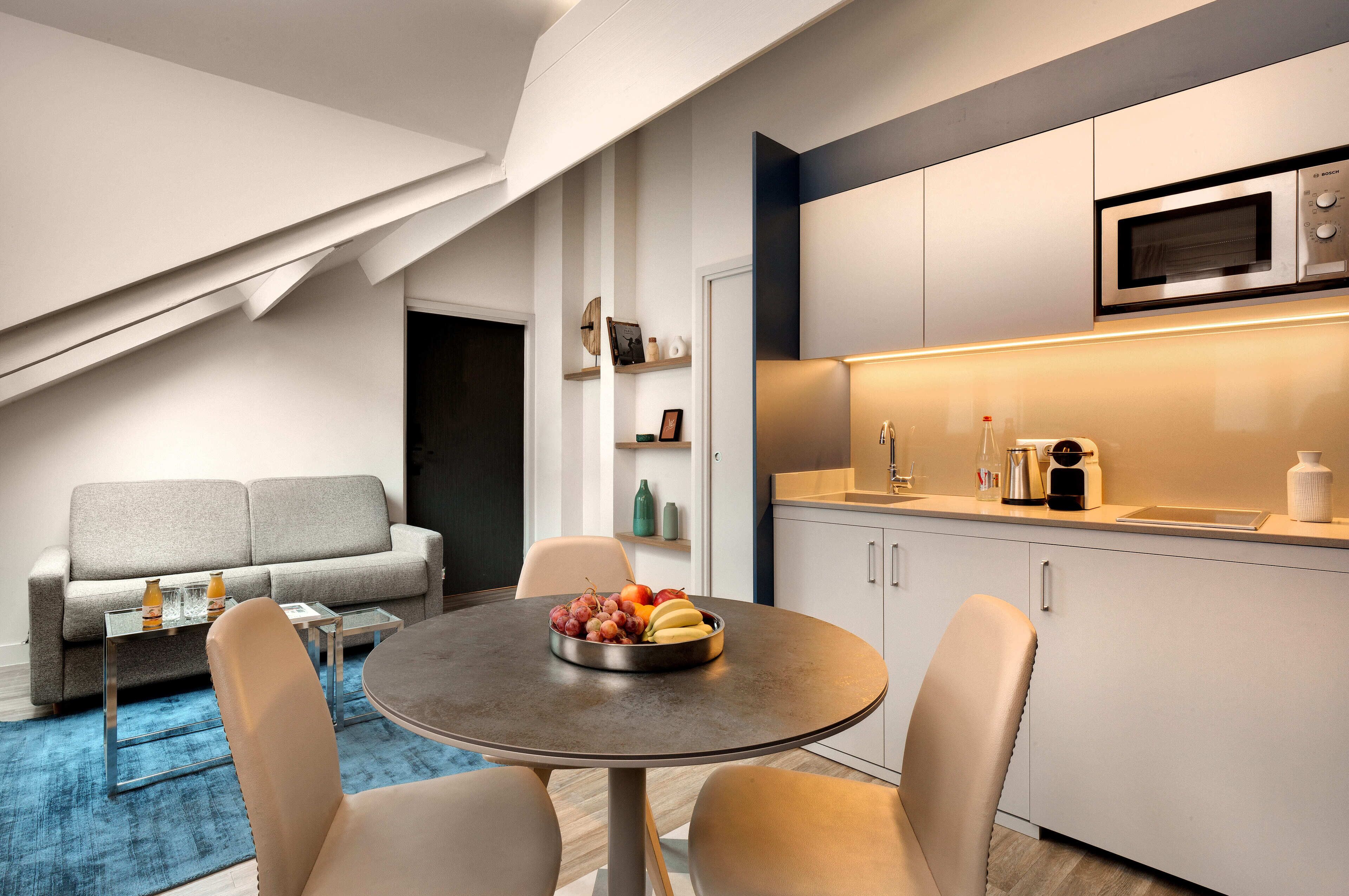 A modern living room with a sofa, coffee table, and a dining table with fruit. A kitchen with a microwave and cabinets is visible.