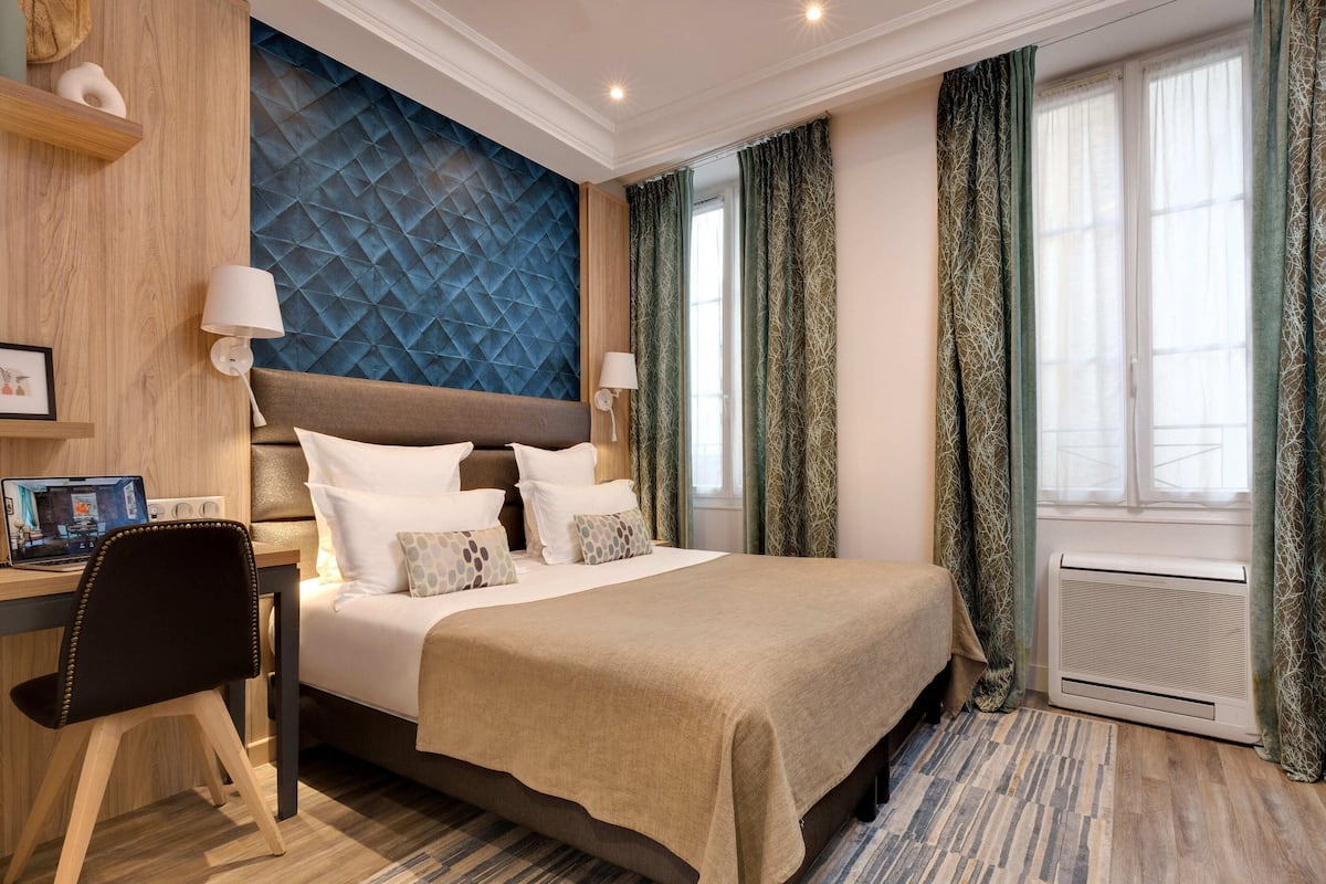 Une chambre d’hôtel avec un lit, un bureau, une chaise et une grande fenêtre avec des rideaux.
