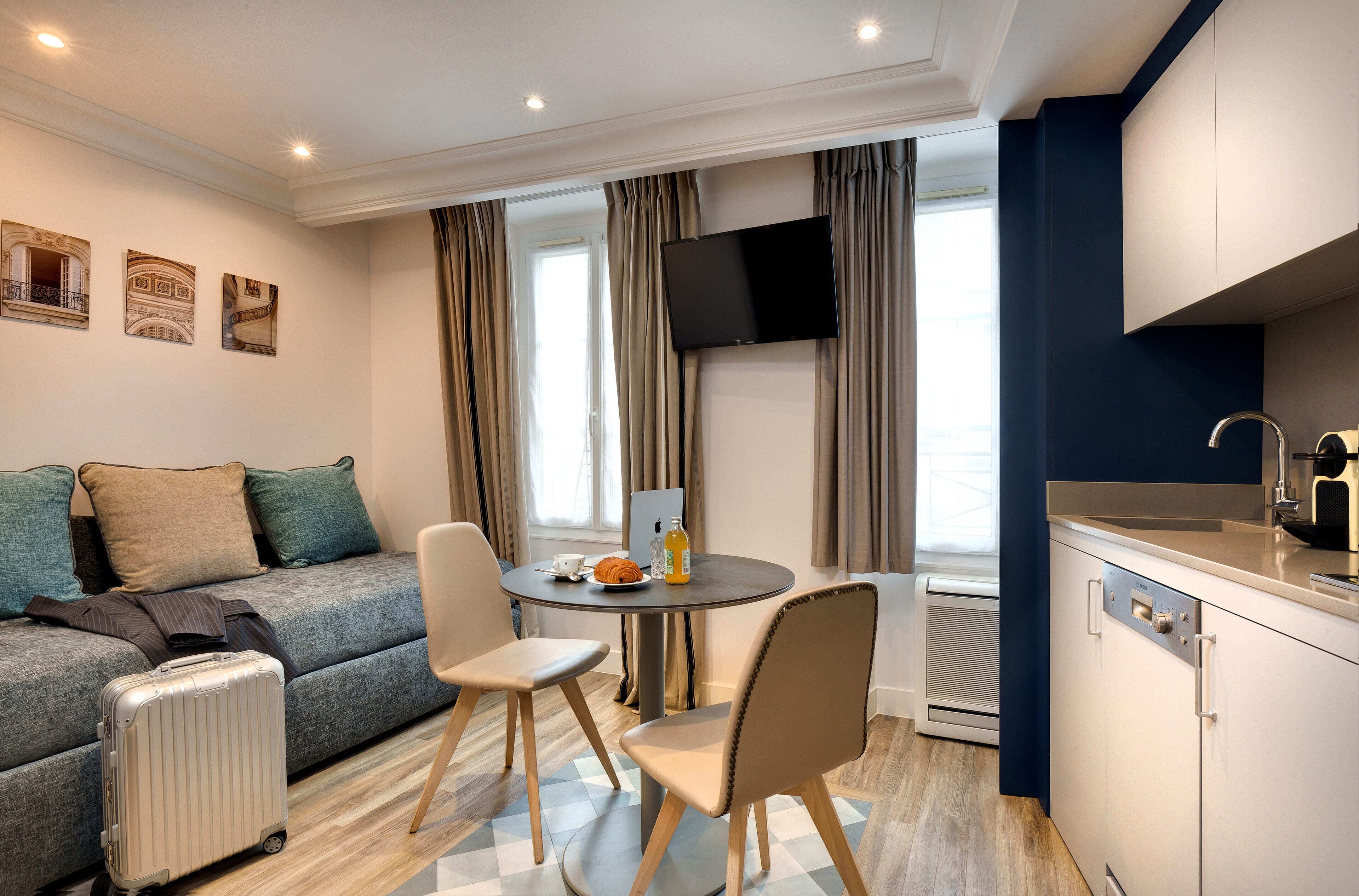 A modern hotel room with a sofa, a dining table, chairs, a TV, and a kitchen area.