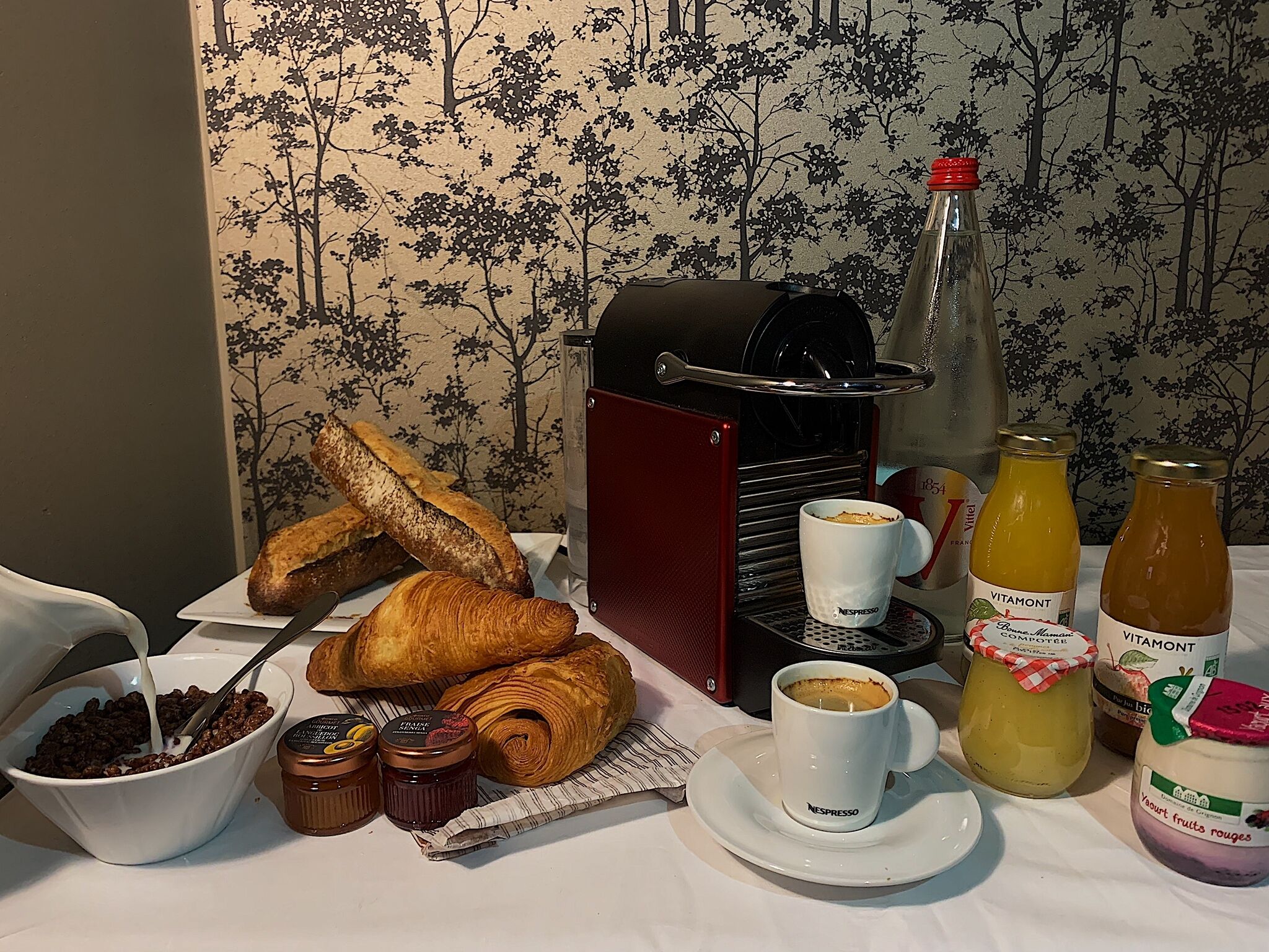 A breakfast setting with croissants, a coffee machine, a cup of coffee, and various condiments.