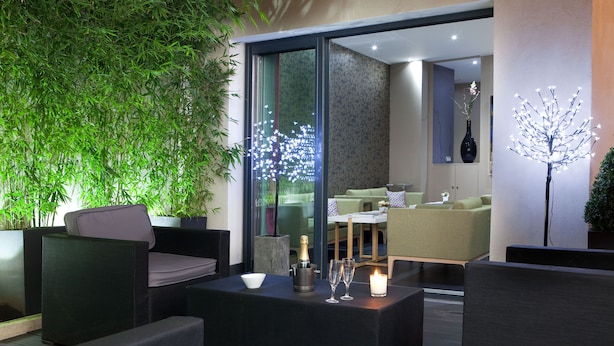 A modern outdoor seating area with a glass partition, green plants, and a decorative light installation. - Serotel Suites (Paris)