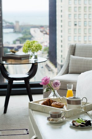 A breakfast tray with croissants, a cup of coffee, and a plate of fruit on a white table in a hotel room with a view of a cityscape.