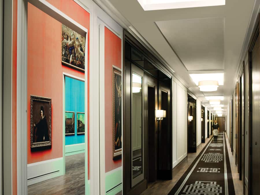 Un pasillo de hotel con alfombra estampada, cuadros enmarcados en las paredes y una fila de puertas.