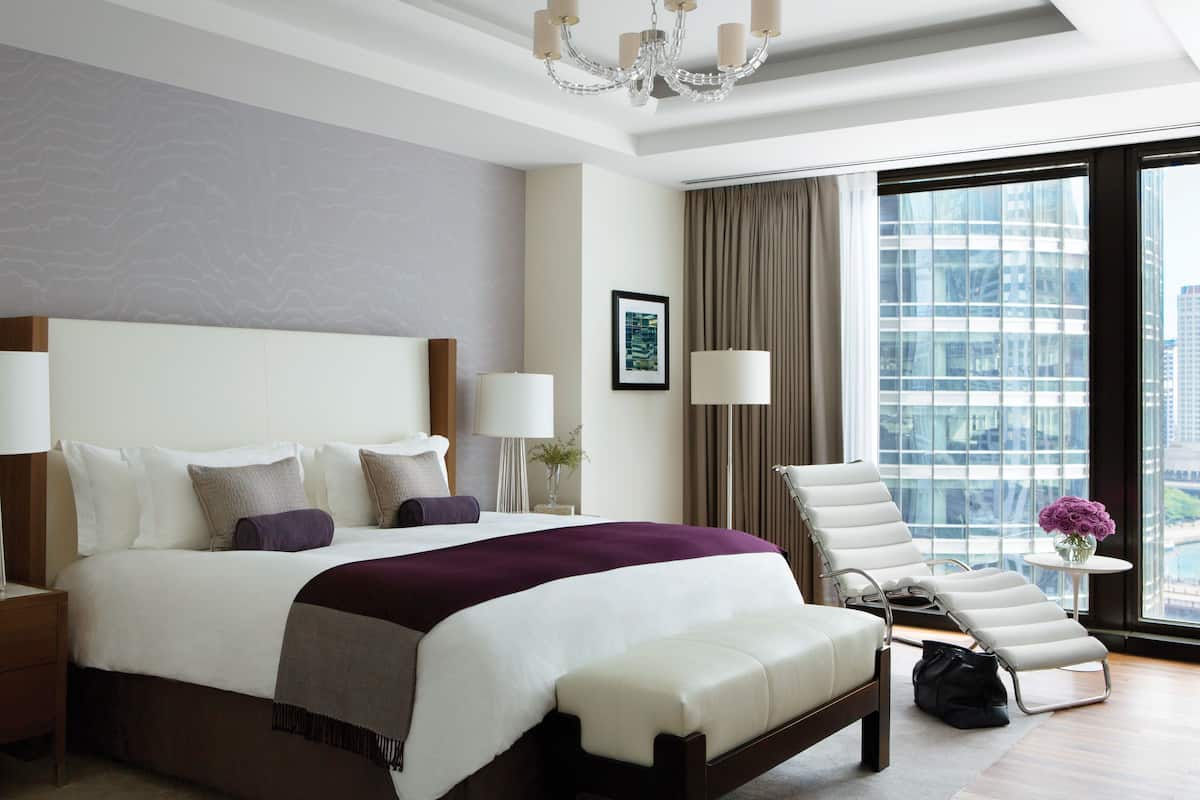 A modern hotel room with a large bed, a sitting area with a chair and a table, a view of the city through large windows, and a chandelier.