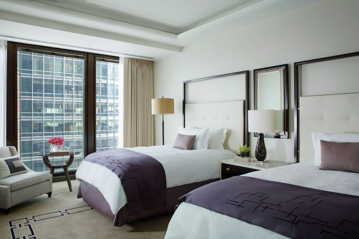 A hotel room with two beds, a sitting area, and a large window with a city view.