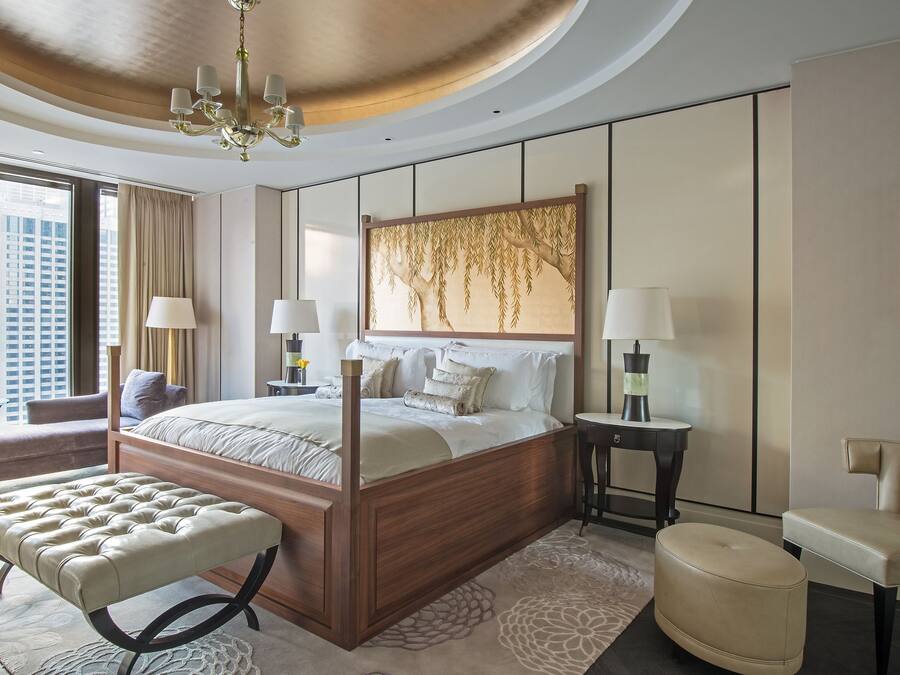 A modern hotel room with a large bed, a sitting area with a bench, a side table, and a view of the city.