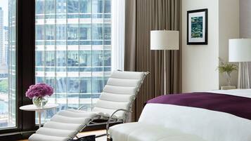 A hotel room with a large window, a bed, a chair, a small table with a vase, and a lamp.