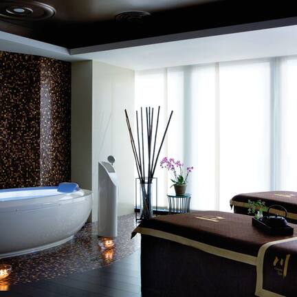 A spa room with a large bathtub, two massage tables, a decorative wall, and a window with natural light.