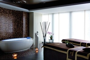 A spa room with a large bathtub, two massage tables, a decorative wall, and a window with natural light.