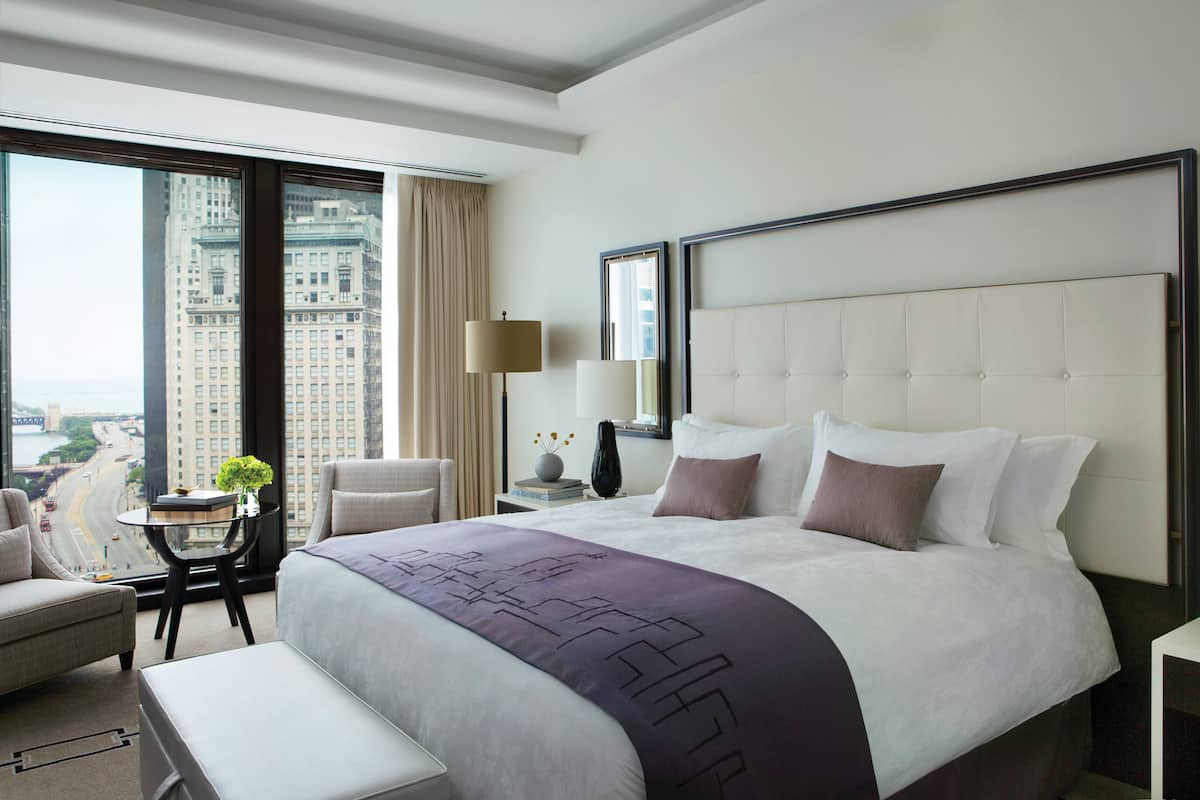 A modern hotel room with a large bed, a view of a cityscape, a small table with a lamp, and a chair.