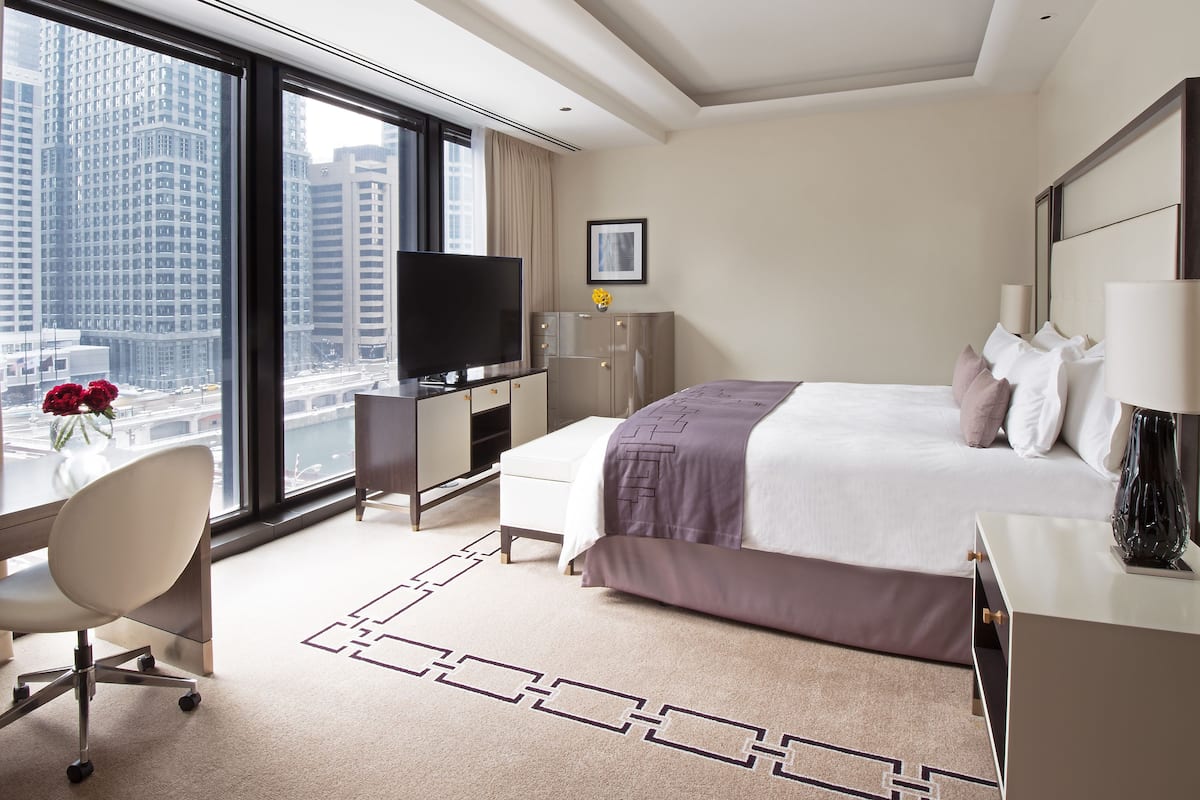 A hotel room with a large bed, a desk with a chair, a TV, and a view of tall buildings.