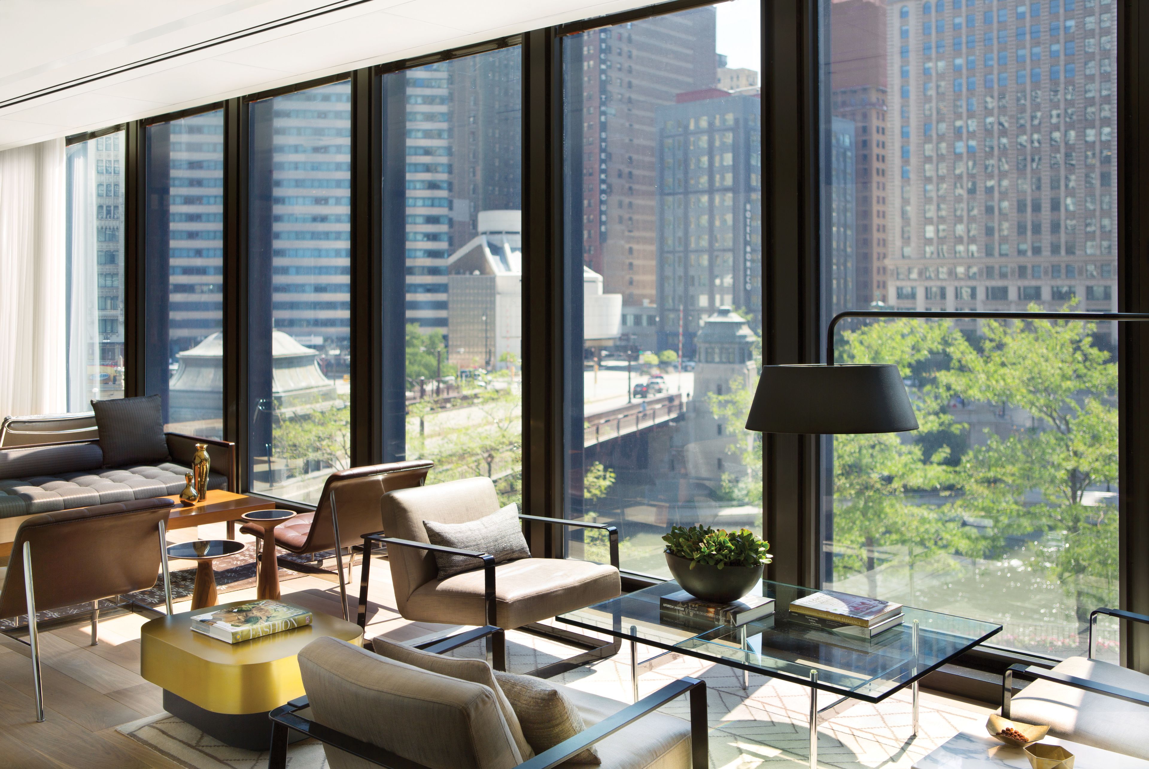 A modern living room with a large city view, featuring a sofa, armchairs, a glass coffee table, and a lamp.