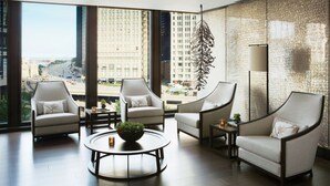Une chambre d’hôtel moderne dotée d’une grande fenêtre donnant sur la ville, de trois fauteuils disposés autour d’une table basse centrale et d’une plante décorative suspendue.