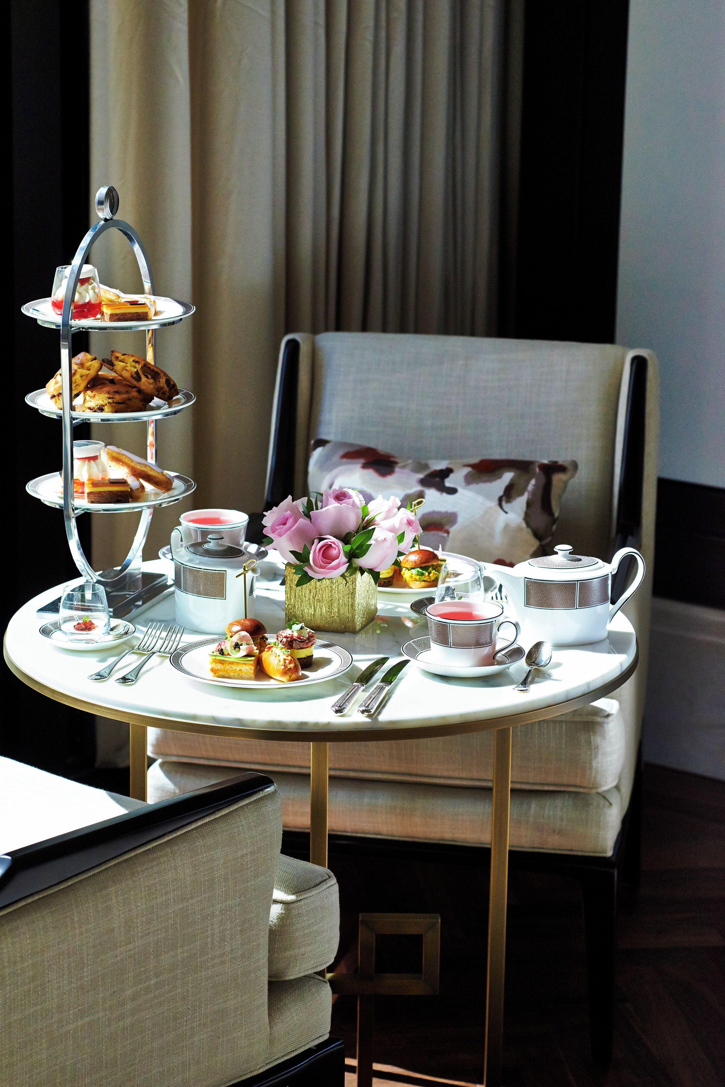 A three-tiered tea service set on a table with various pastries and a vase of flowers, placed in a room with a chair and curtains in the background.