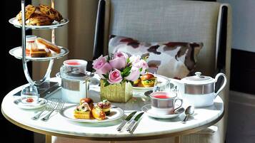 A three-tiered tea service set on a table with various pastries and a vase of flowers, placed in a room with a chair and curtains in the background.