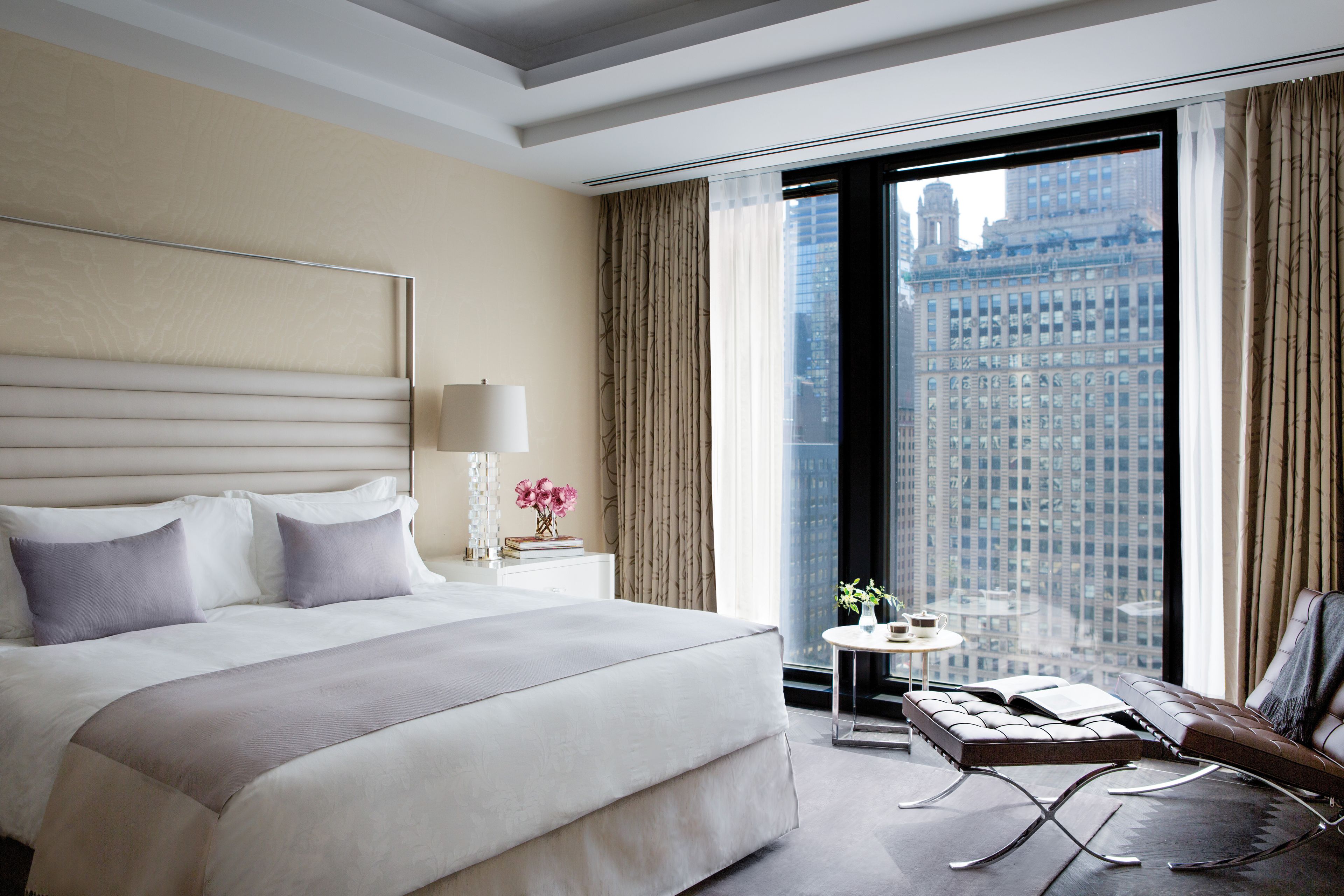 Une chambre d’hôtel moderne avec un grand lit, une petite table avec une lampe, une chaise et une vue sur le paysage urbain par une grande fenêtre.
