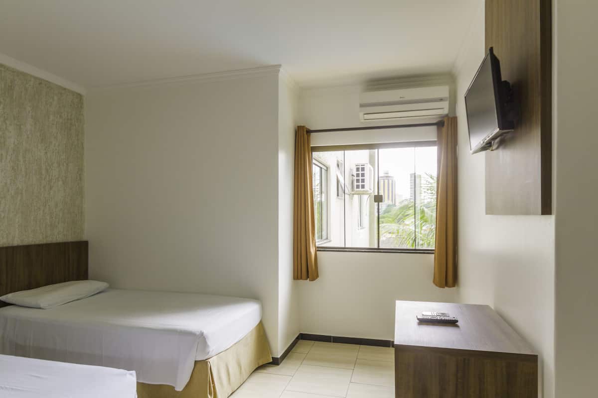 A hotel room with two single beds, a small desk with a remote control, a TV mounted on the wall, and a window with curtains.