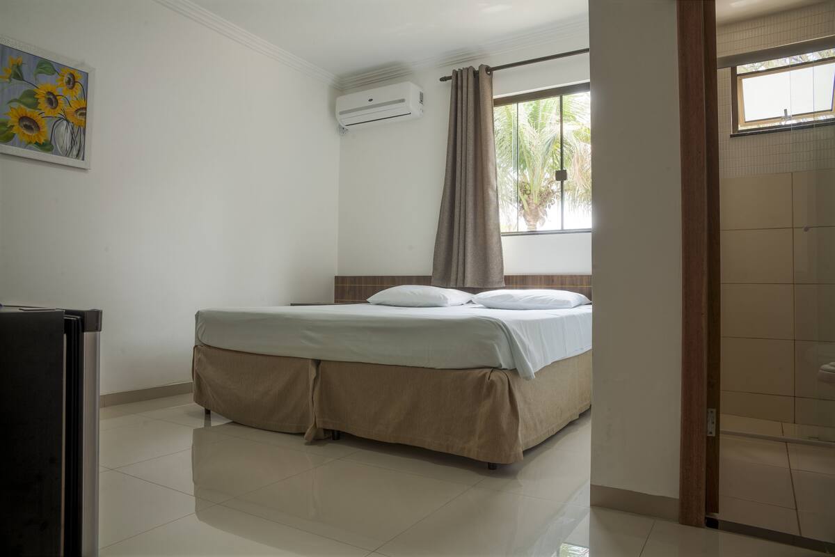 A hotel room with a bed, a window with curtains, a wall-mounted air conditioner, and a painting of sunflowers.