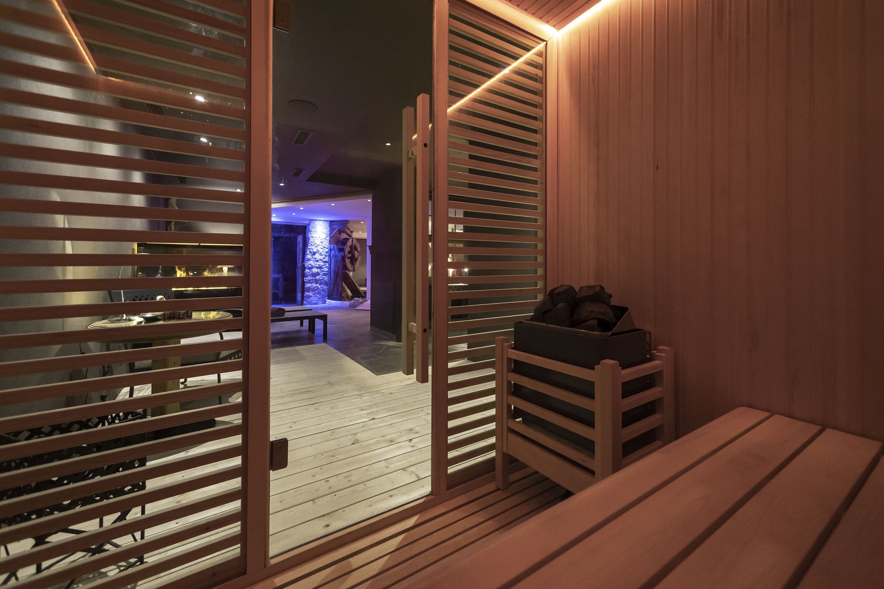 A wooden interior with a bench and a shoe rack, a wooden door with horizontal slats, and a glimpse of a room with a blue-lit wall and a bench in the background.