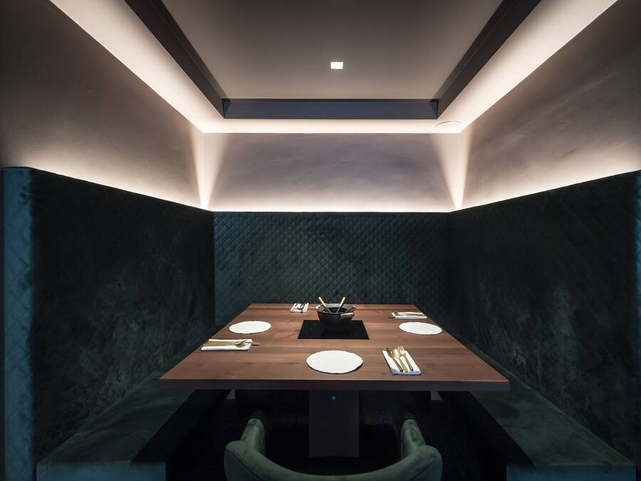 Uma sala de jantar moderna com uma mesa retangular, paredes texturizadas escuras e iluminação embutida no teto.