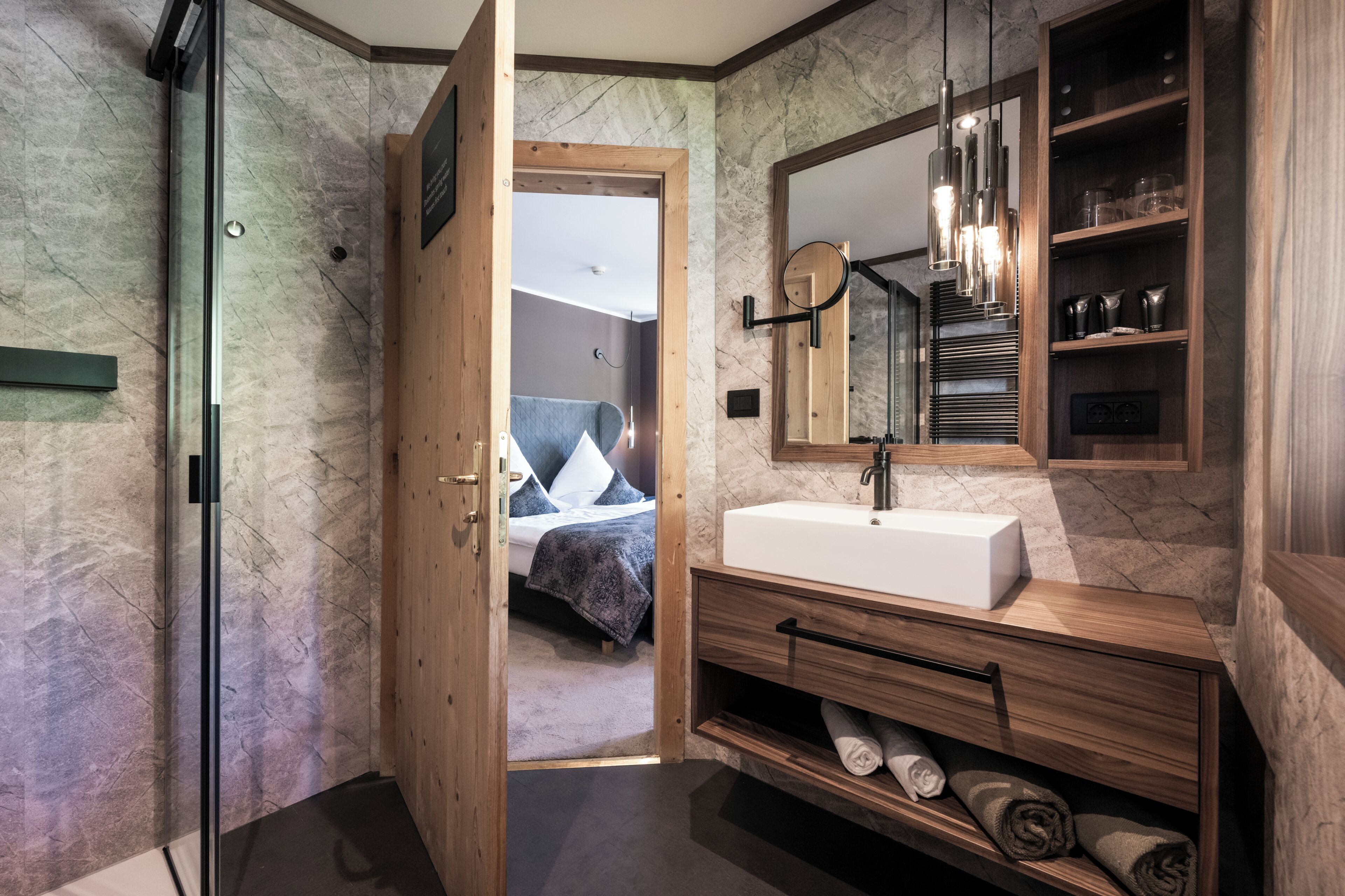 A modern bathroom with a marble wall, a wooden vanity, a large mirror, and a built-in shower.