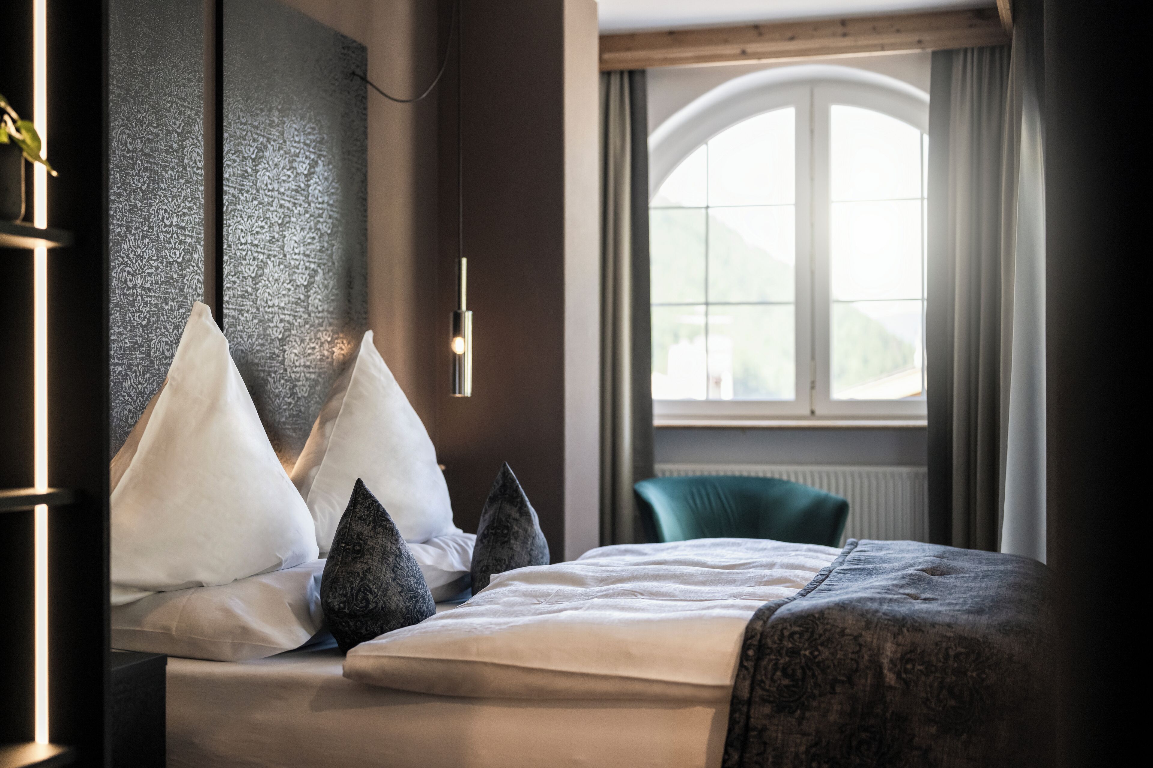 A neatly made bed with white and dark pillows, a bedside table with a lamp, and a view of a window with curtains.