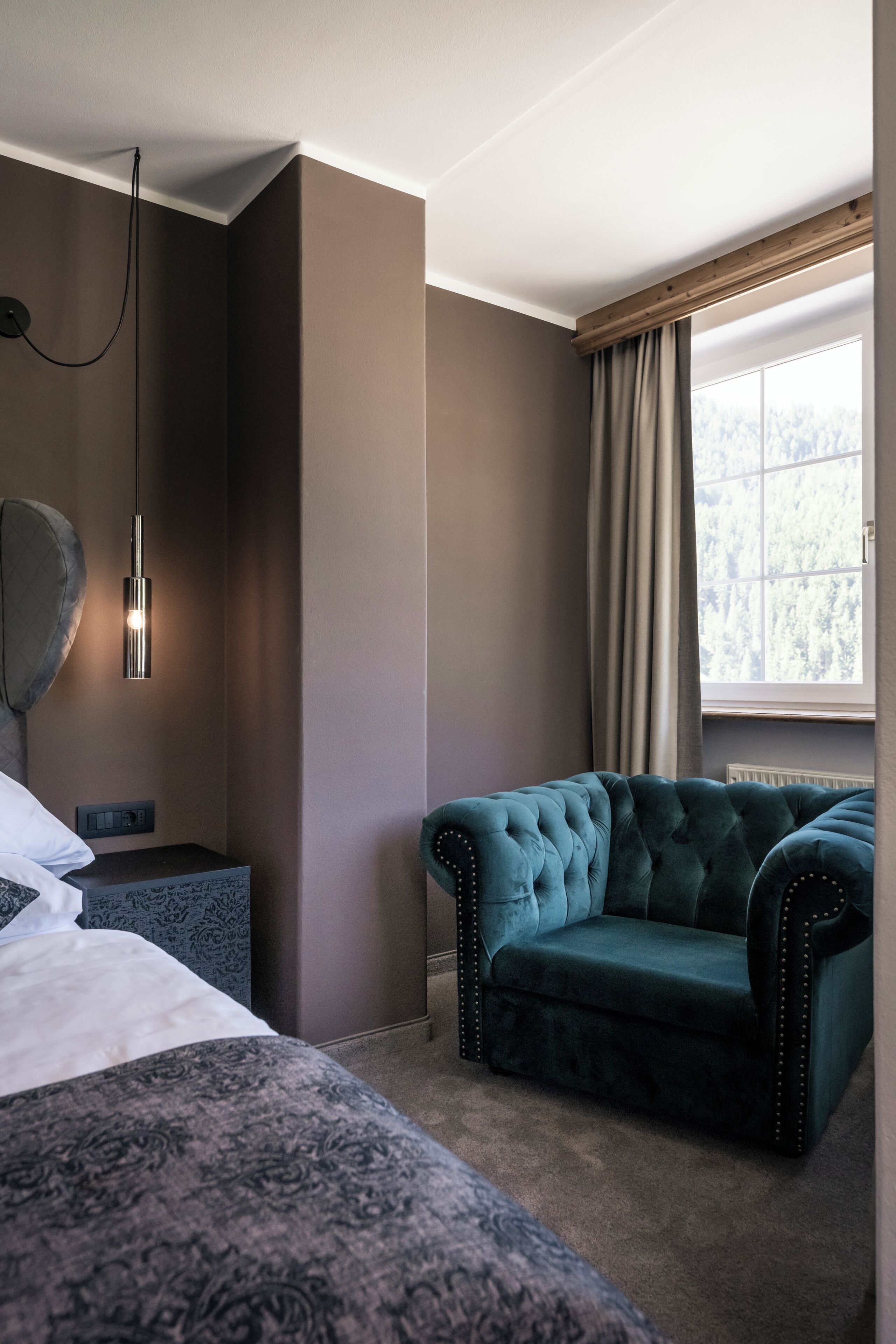 A hotel room with a large bed, a tufted armchair, a bedside table, a lamp, and a window with curtains.
