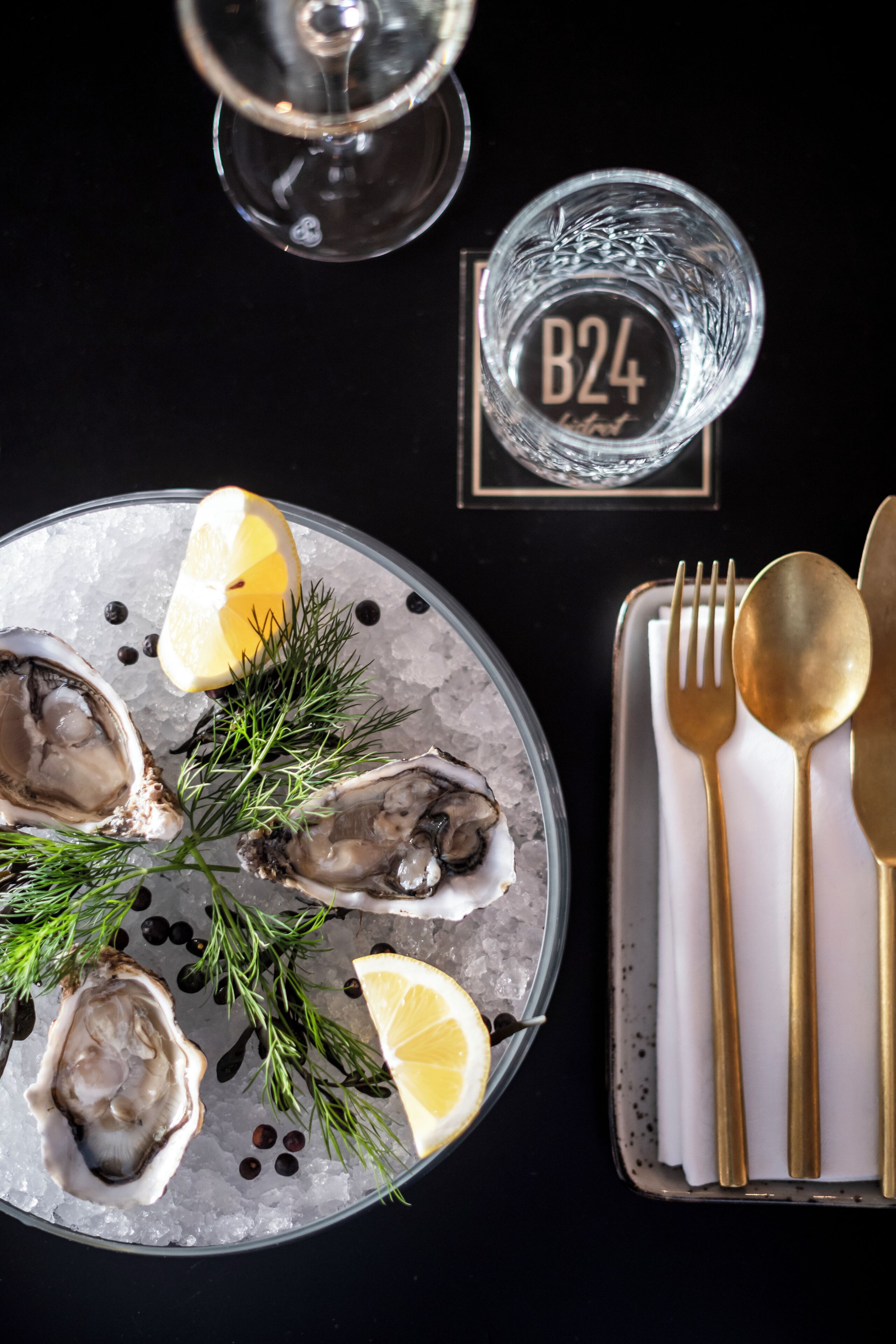 A plate of oysters with lemon and herbs, a glass of water, and gold cutlery on a black table.