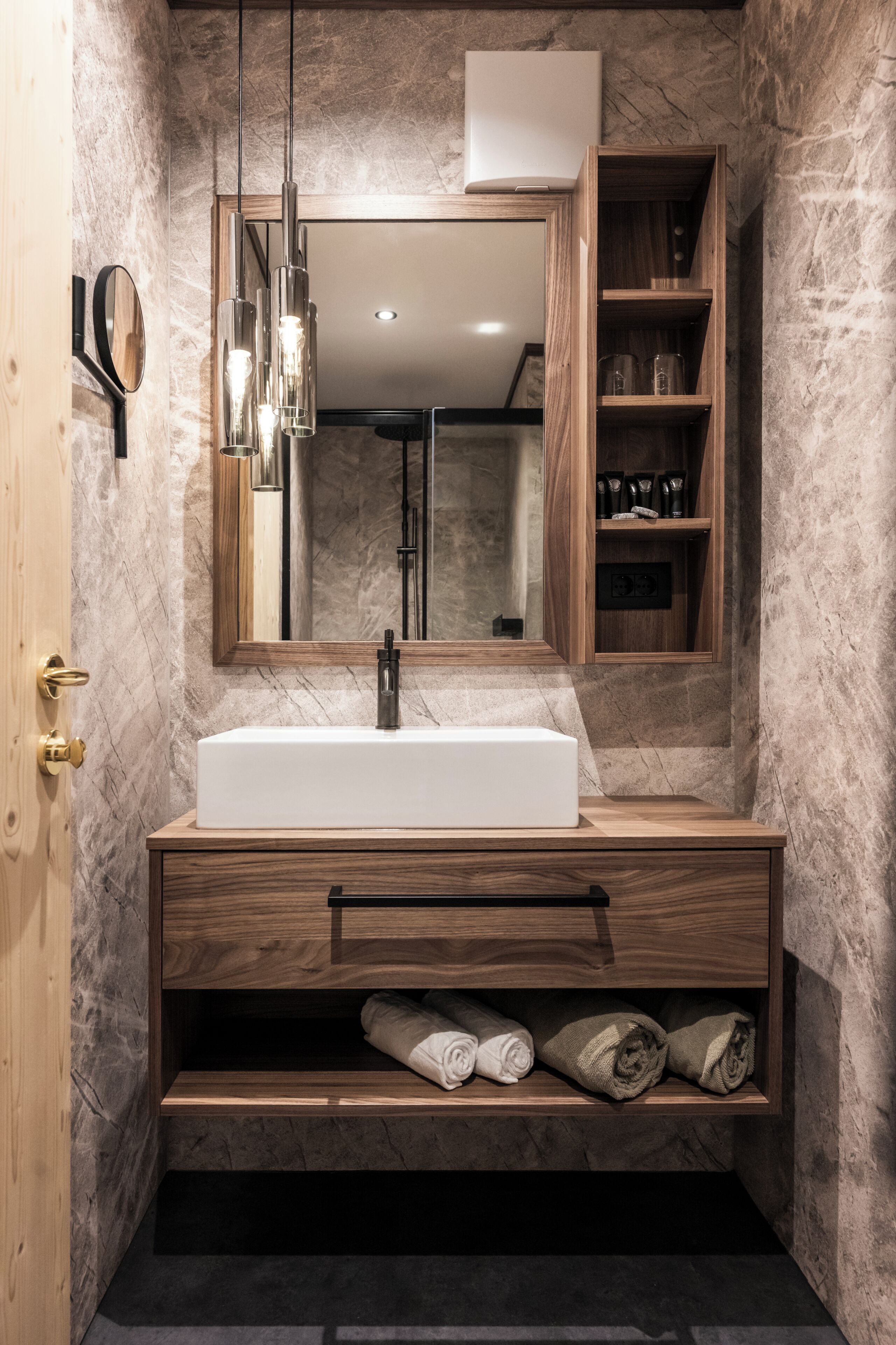 Un baño moderno con pared de mármol, tocador de madera, espejo grande y estante empotrado con artículos de aseo.