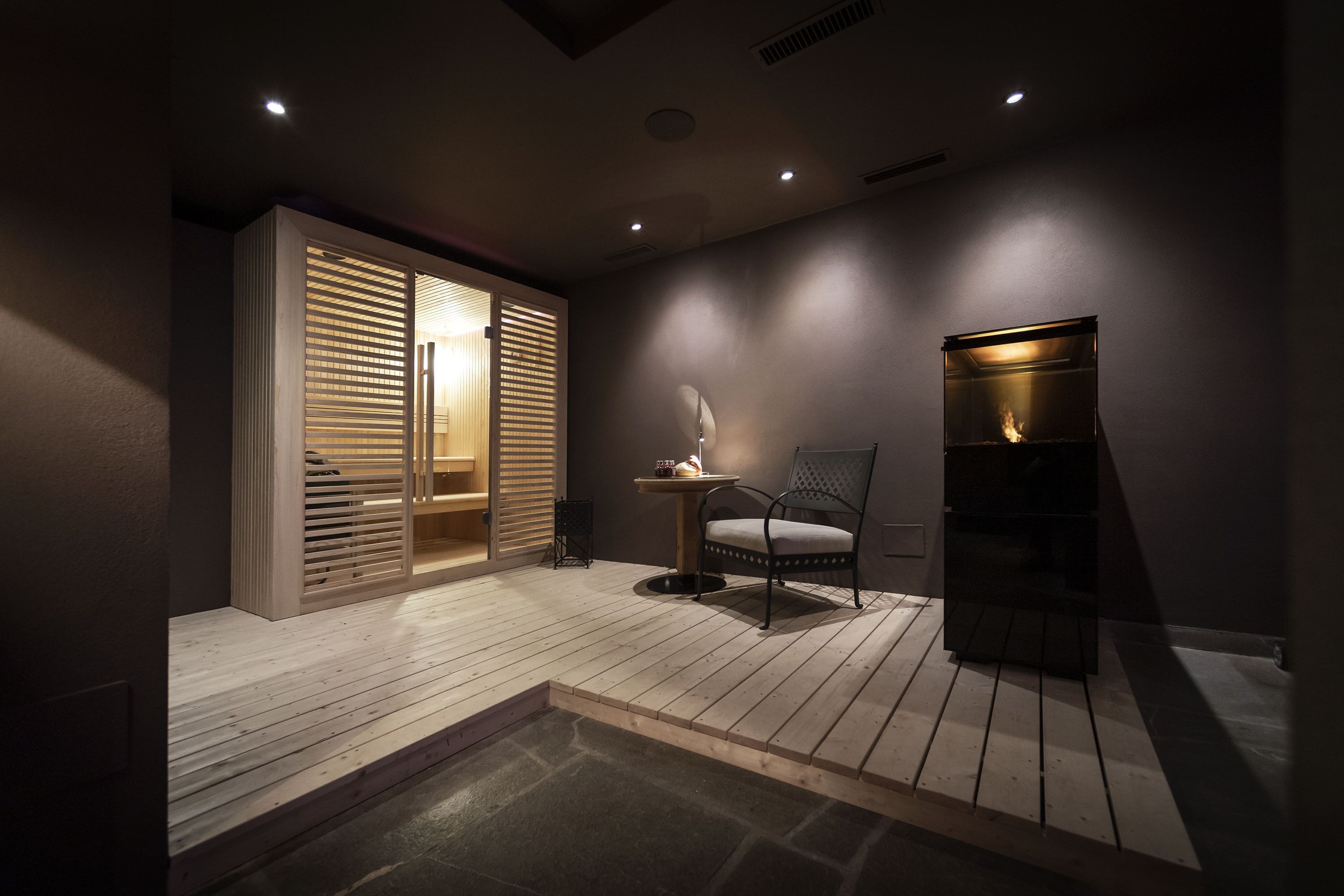 Una habitación moderna con poca luz, suelo de madera, una mesa pequeña con una lámpara, una silla y una estantería empotrada.