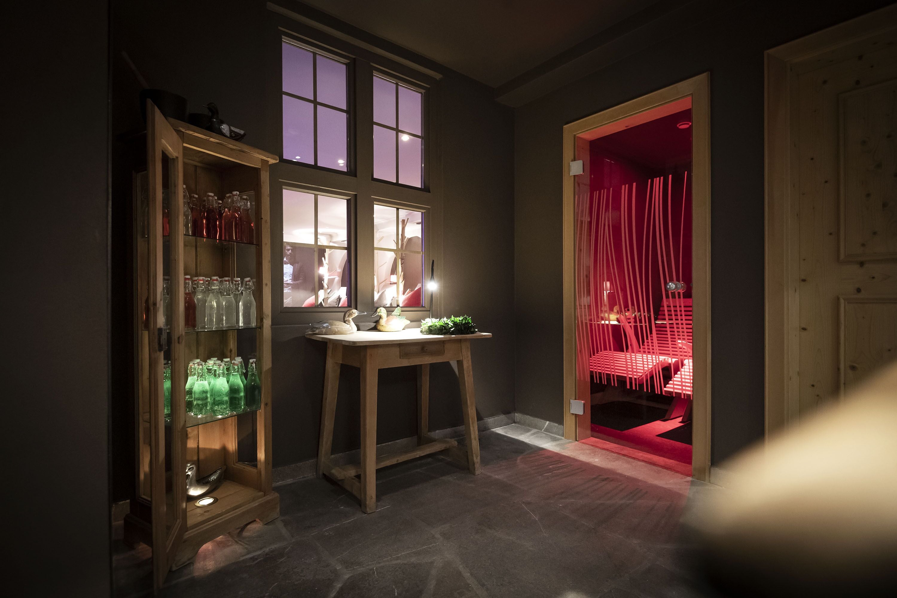 A dimly lit room with a wooden cabinet containing glass bottles, a table with a lamp, and a red-lit doorway leading to another room.