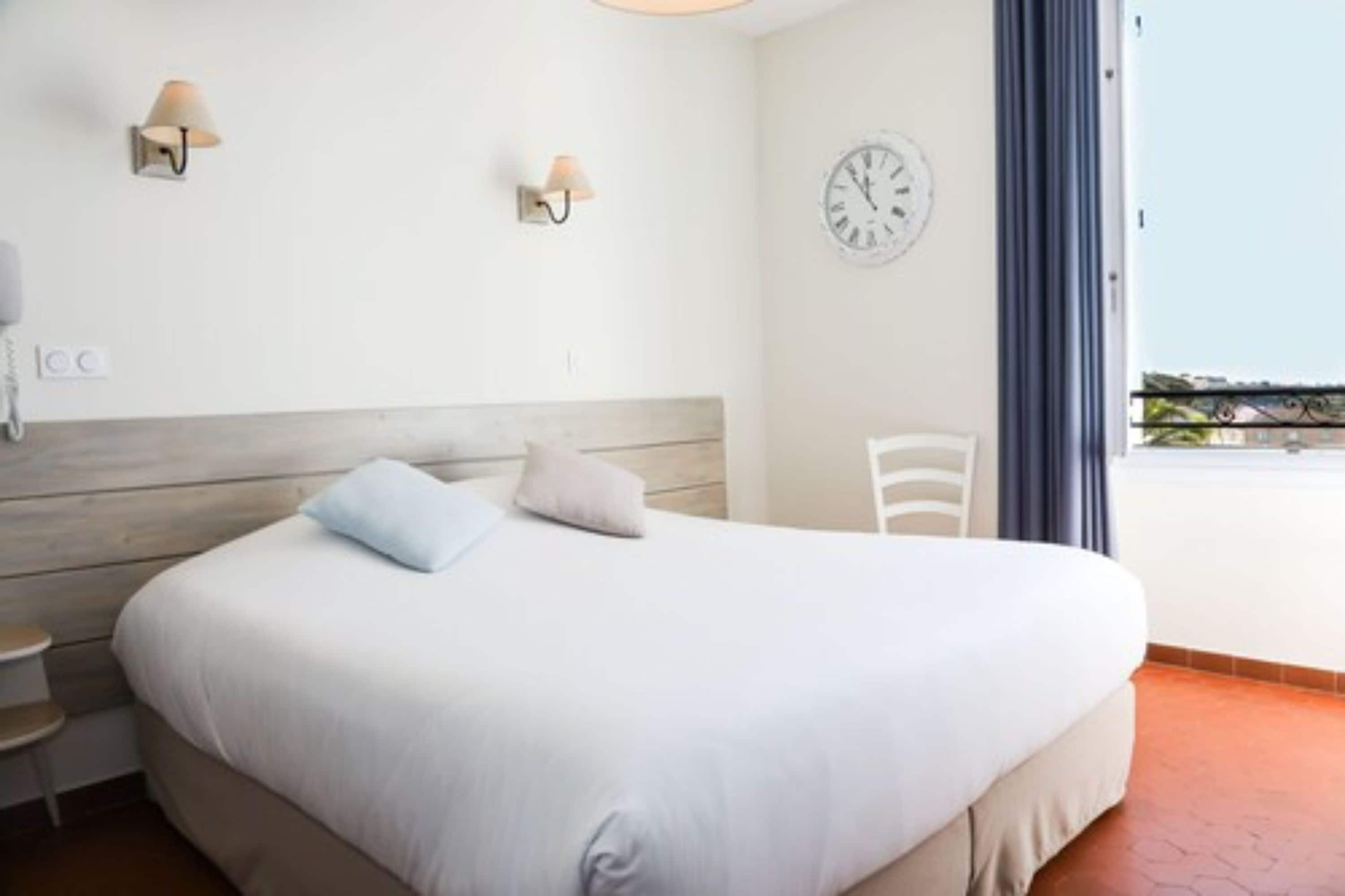 Hotellihuone, jossa on sänky, kaksi seinälle asennettua lamppua, kello ja näkymä ulos.