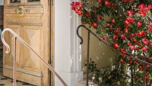 Una puerta de madera con ornamentación metálica intrincada, una pared blanca y una escalera que lleva a un balcón con una planta floreciente roja.